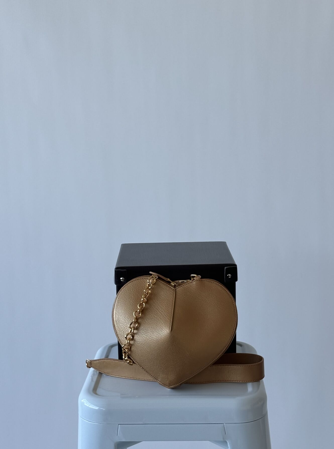 An ALAÏA Le Coeur leather shoulder bag by EXCHANGE, featuring a gold heart shape and chain strap, rests on a black box atop a white stool against a plain light gray background. This is the FULL SET.