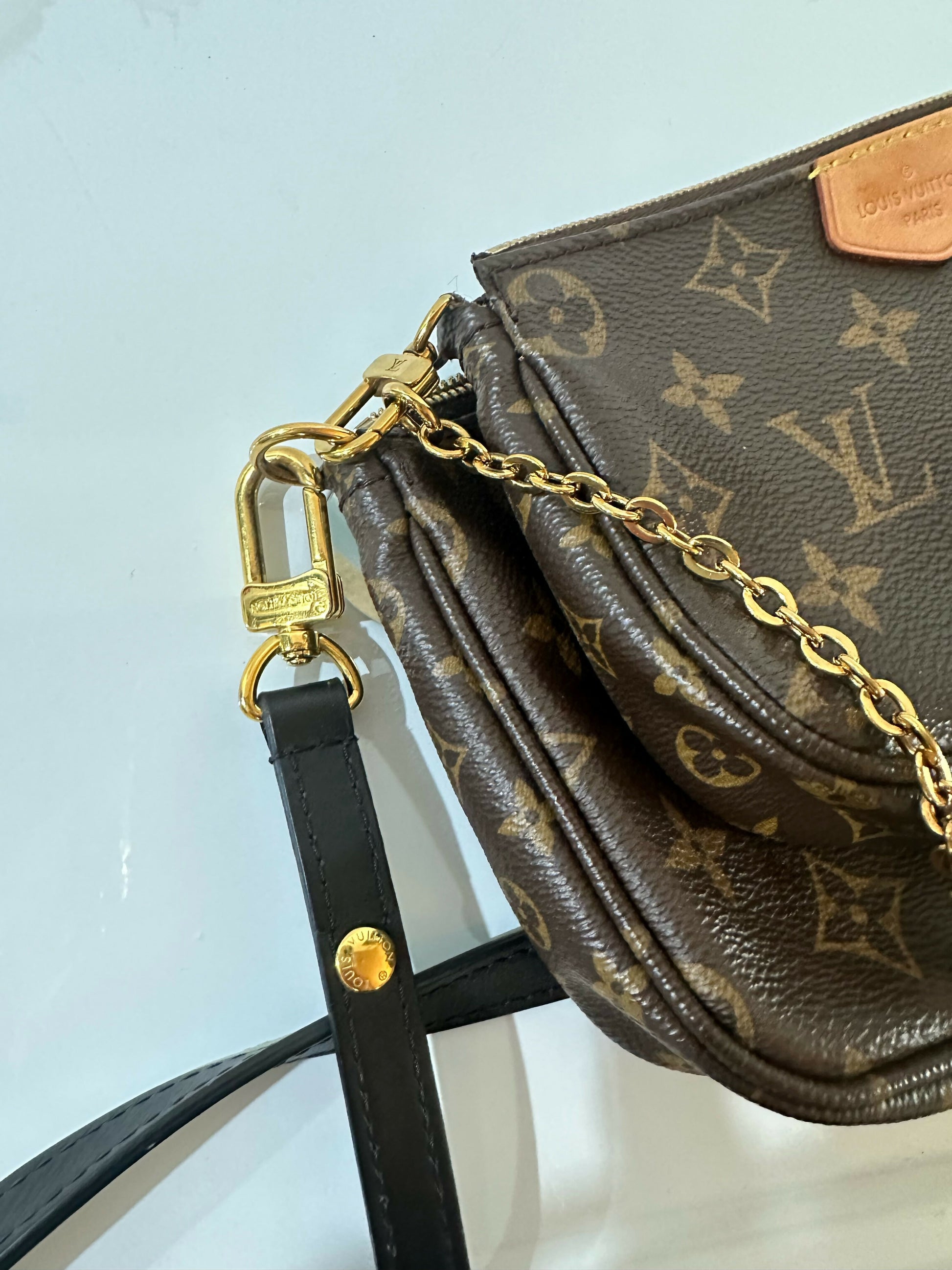 Close-up of the Louis Vuitton Multi Pochette - Full Set by EXCHANGE, featuring its gold chain, branded clasp, and black leather strap with a gold button against a light background.