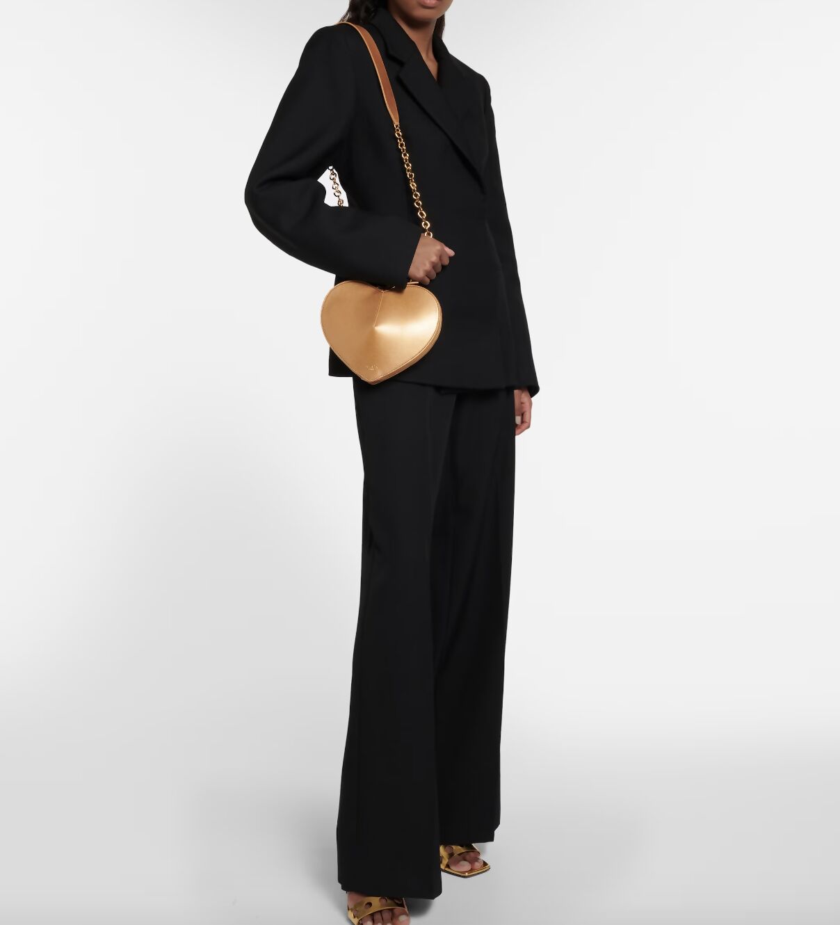 Wearing a black suit with flared trousers, someone holds the EXCHANGE ALAÏA Le Coeur leather shoulder bag – FULL SET, featuring a chain strap, paired with gold open-toe heels, set against a plain white background.