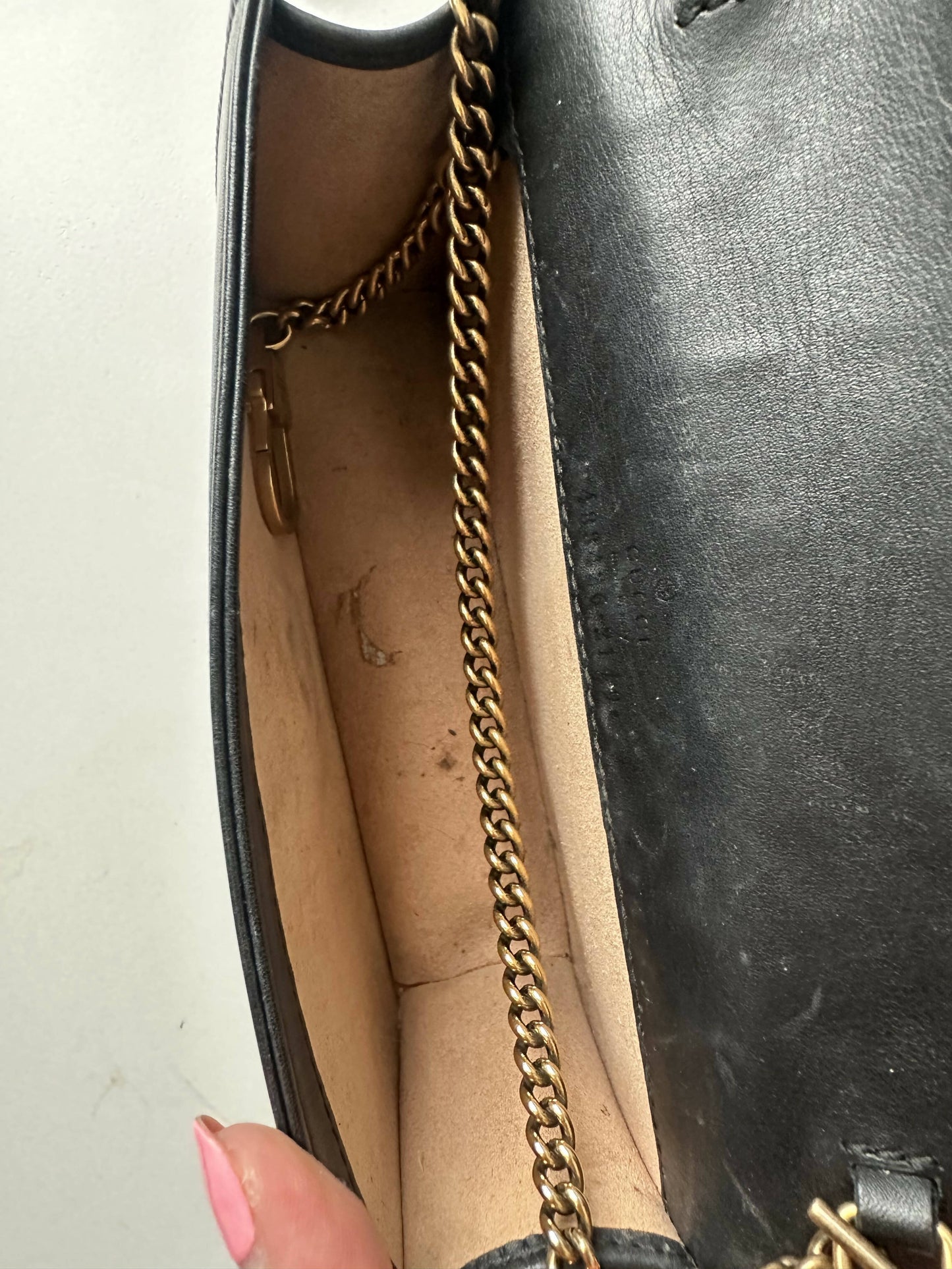 Open black Gucci Marmont Butterfly Bag by EXCHANGE with beige interior, gold chain strap, and some visible dirt or stains inside. A hand with long pink nails holds the purse open.