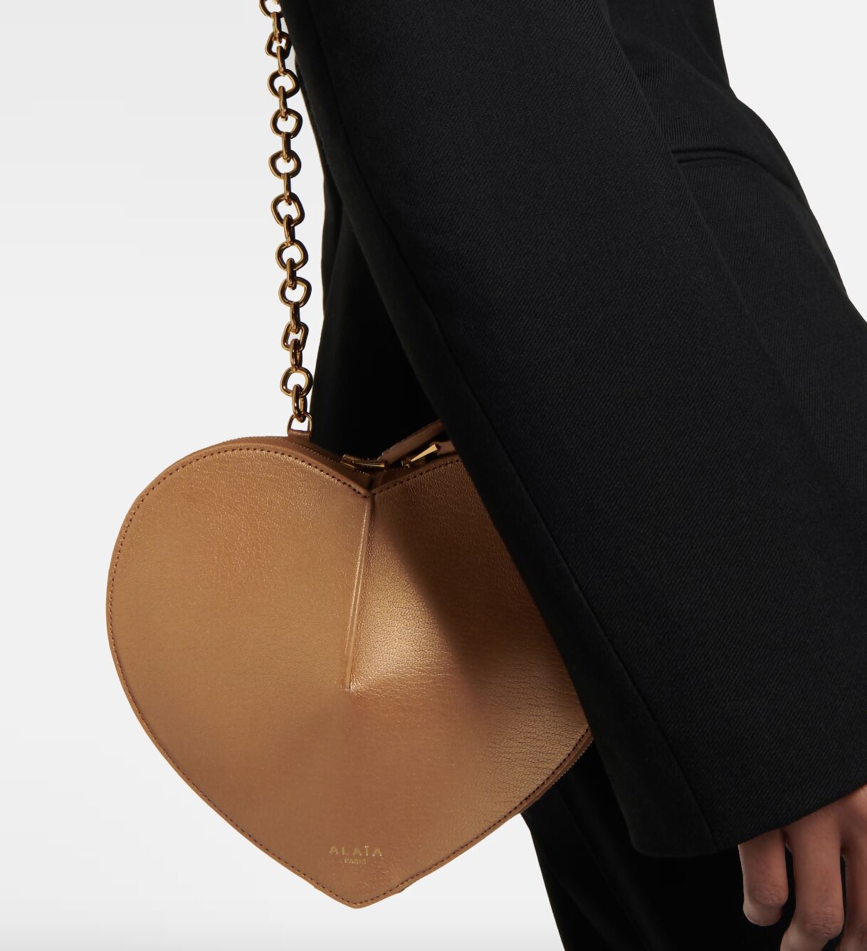 A person in black carries the EXCHANGE ALAÏA Le Coeur leather shoulder bag - FULL SET, a tan heart-shaped handbag with a gold chain strap and an embossed logo near the bottom.
