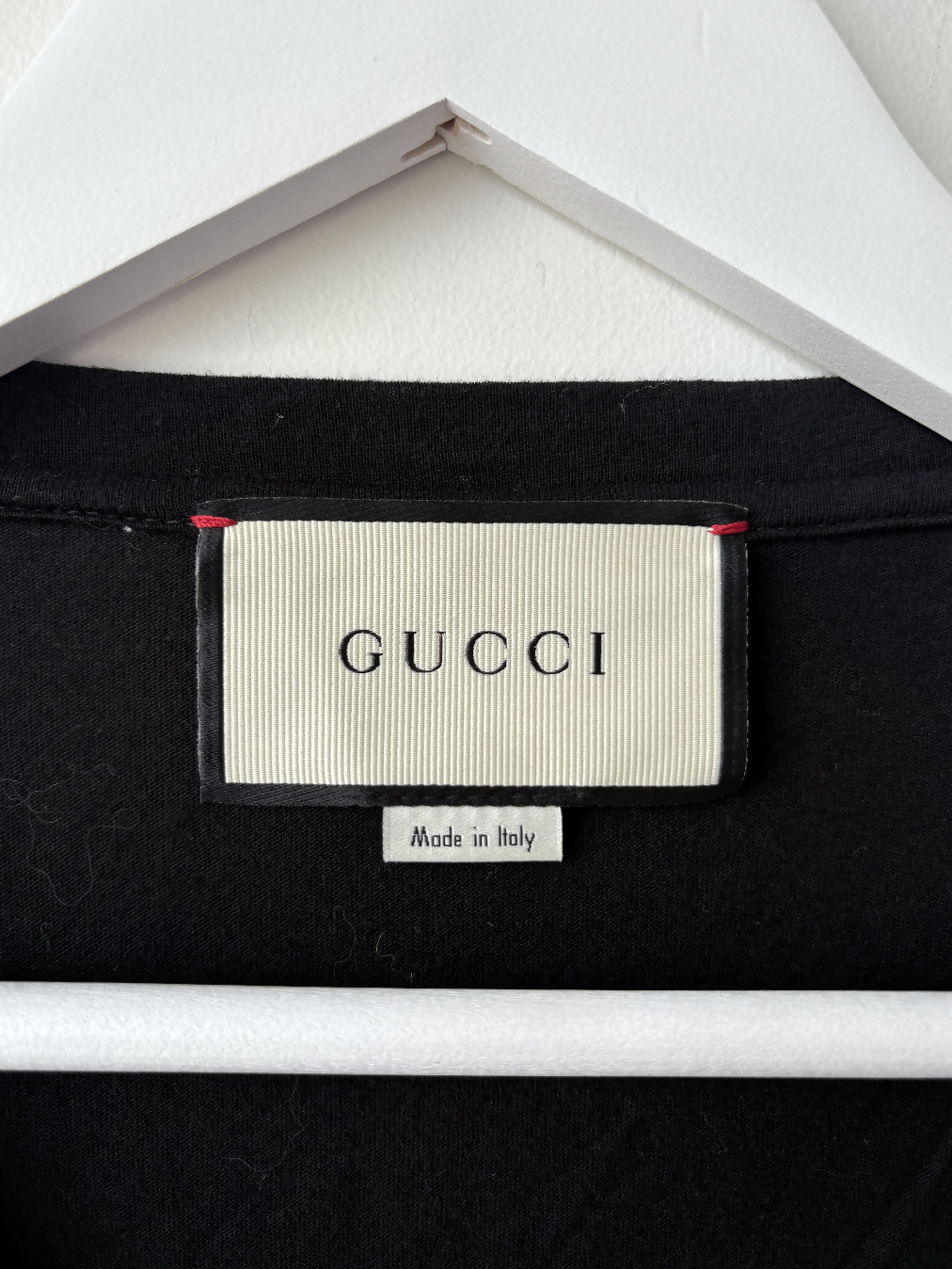 A close-up of the Gucci Black L'Aveugle Par Amour T-Shirt tag by EXCHANGE STORE, featuring the classic logo in black on white and a "Made in Italy" label, stylishly displayed on a white hanger.