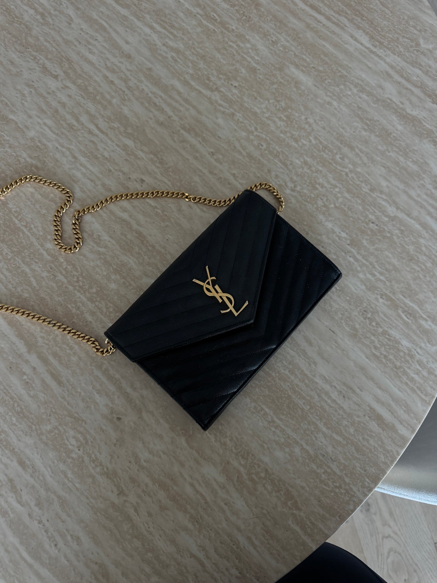 A Saint Laurent Classic Cassandre chain wallet in grain de poudre leather by EXCHANGE STORE, black with gold chain strap and logo, rests on a light-colored, textured tabletop.