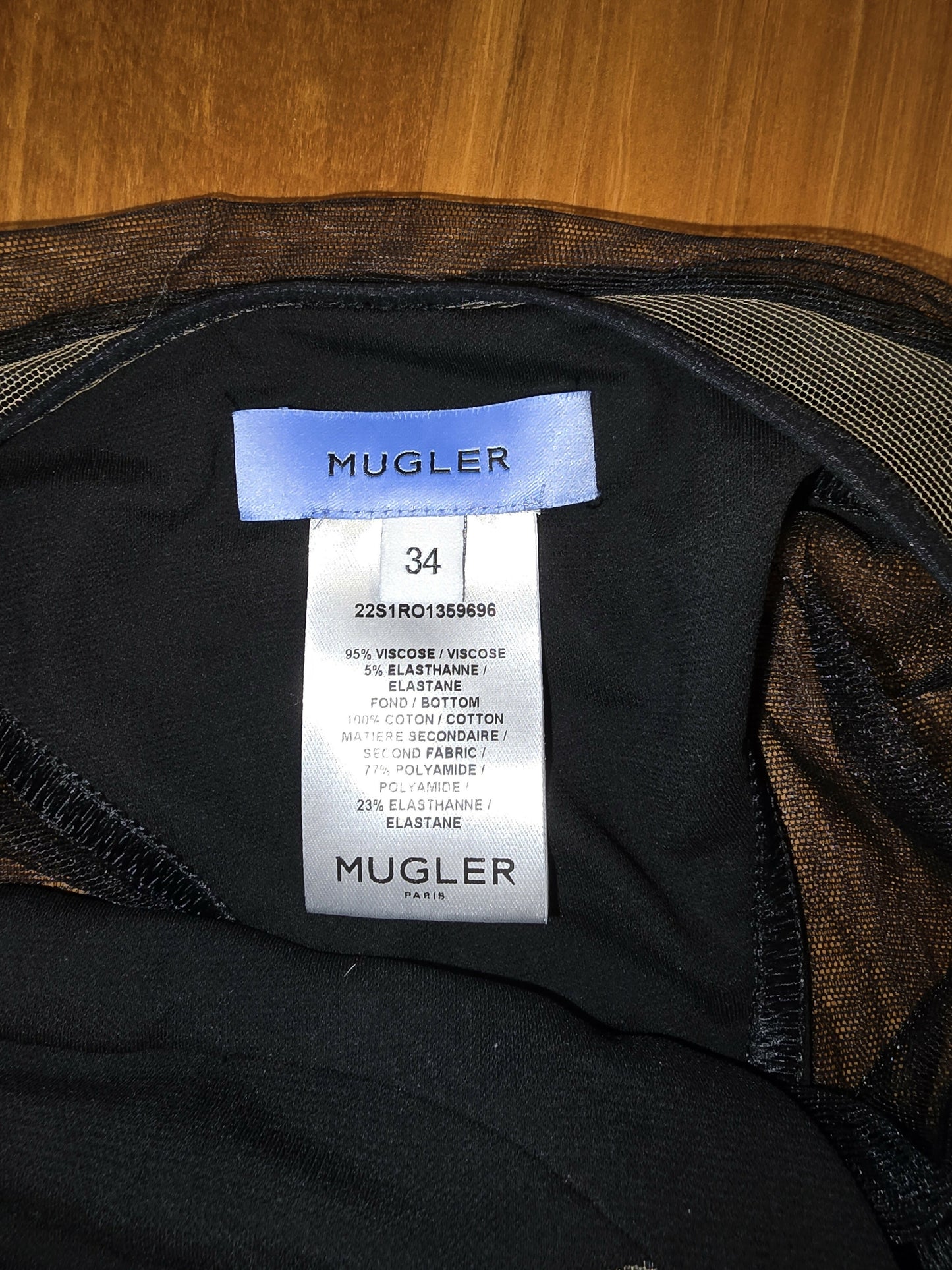 A close-up of a shiny white label inside a black EXCHANGE MUGLER cutout stretch-jersey midi dress, size 34. The tag lists fabric details, care instructions, and states France as the origin. Dress looks like new on a wooden background.