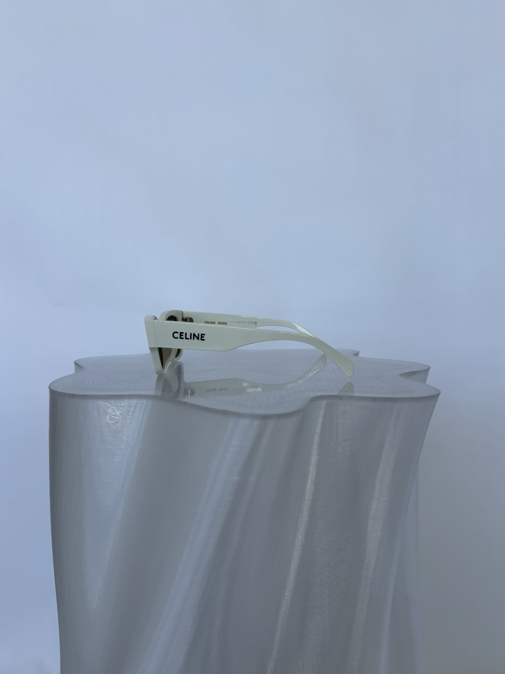 A pair of Celine Beige Sunglasses by EXCHANGE STORE is displayed on a translucent, wavy-edged pedestal against a light blue background.