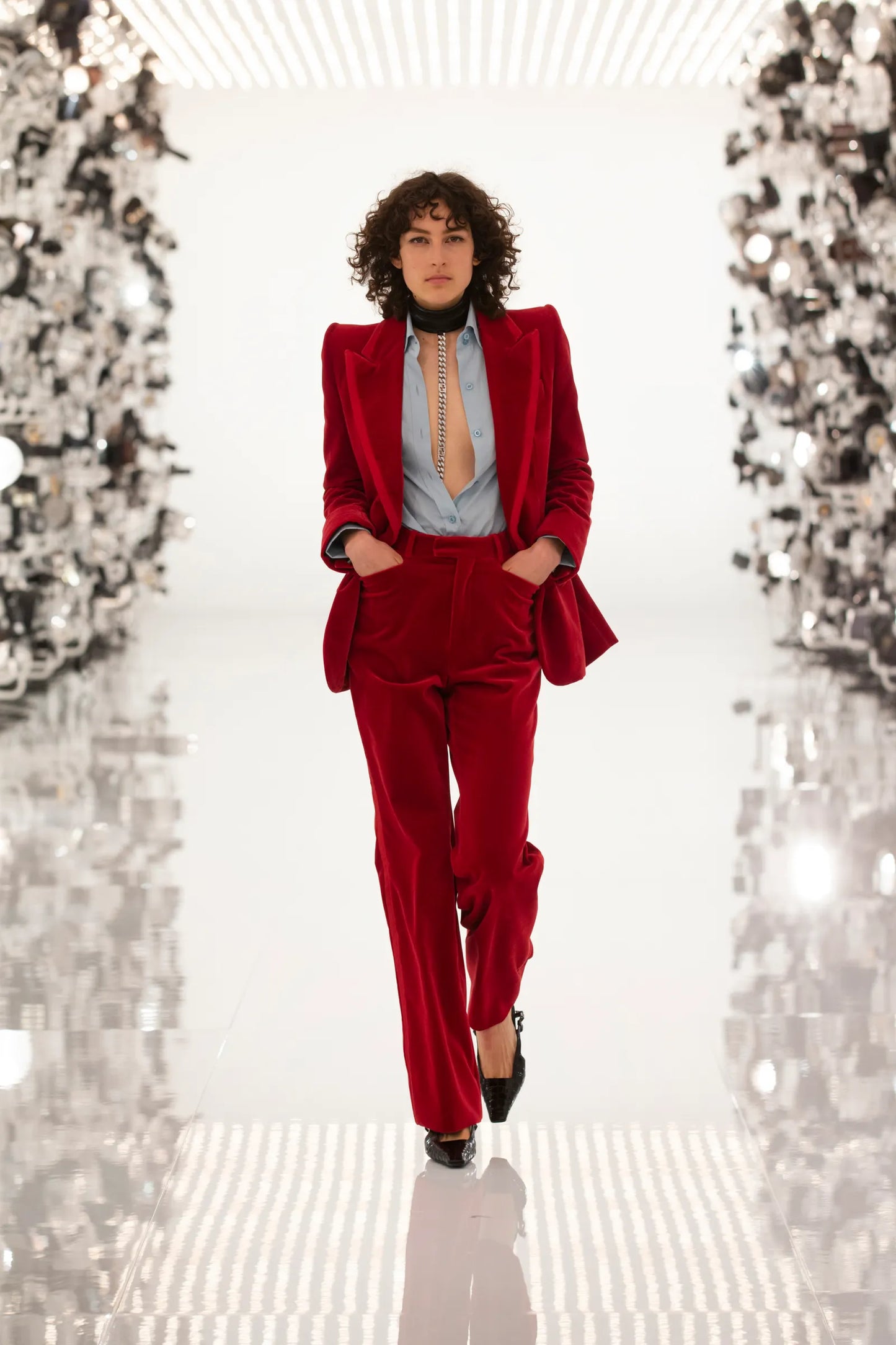 A model struts on a glossy runway in the EXCHANGE Tom Ford Red Velvet Gucci Suit, paired with a light blue unbuttoned shirt, black choker, and heels. The scene is illuminated by bright lights and reflective silver decor.