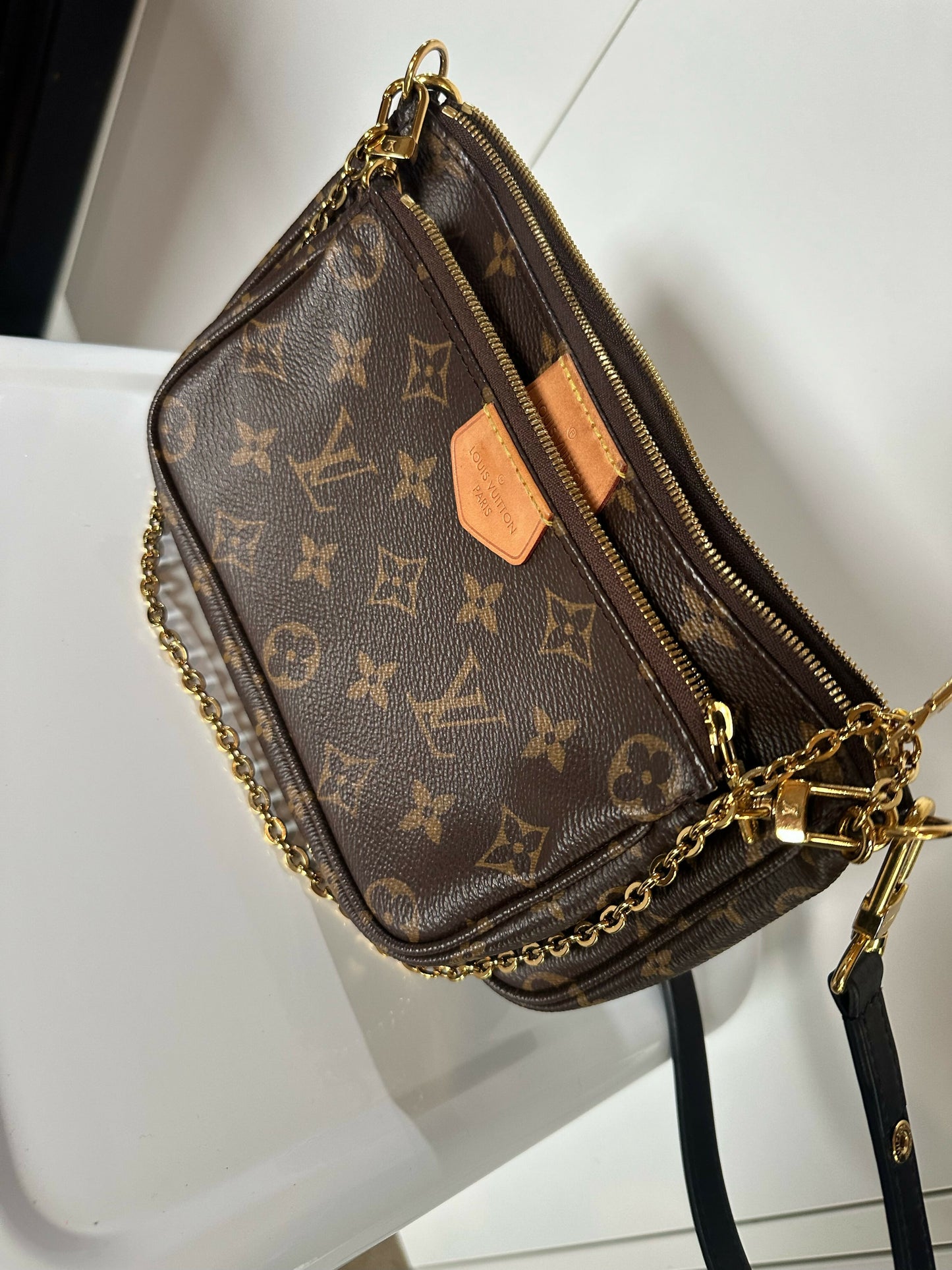 A brown EXCHANGE Louis Vuitton Multi Pochette - Full Set in Monogram canvas with gold zipper and chain strap, tan leather tags, and matching hardware accents rests on a white surface.