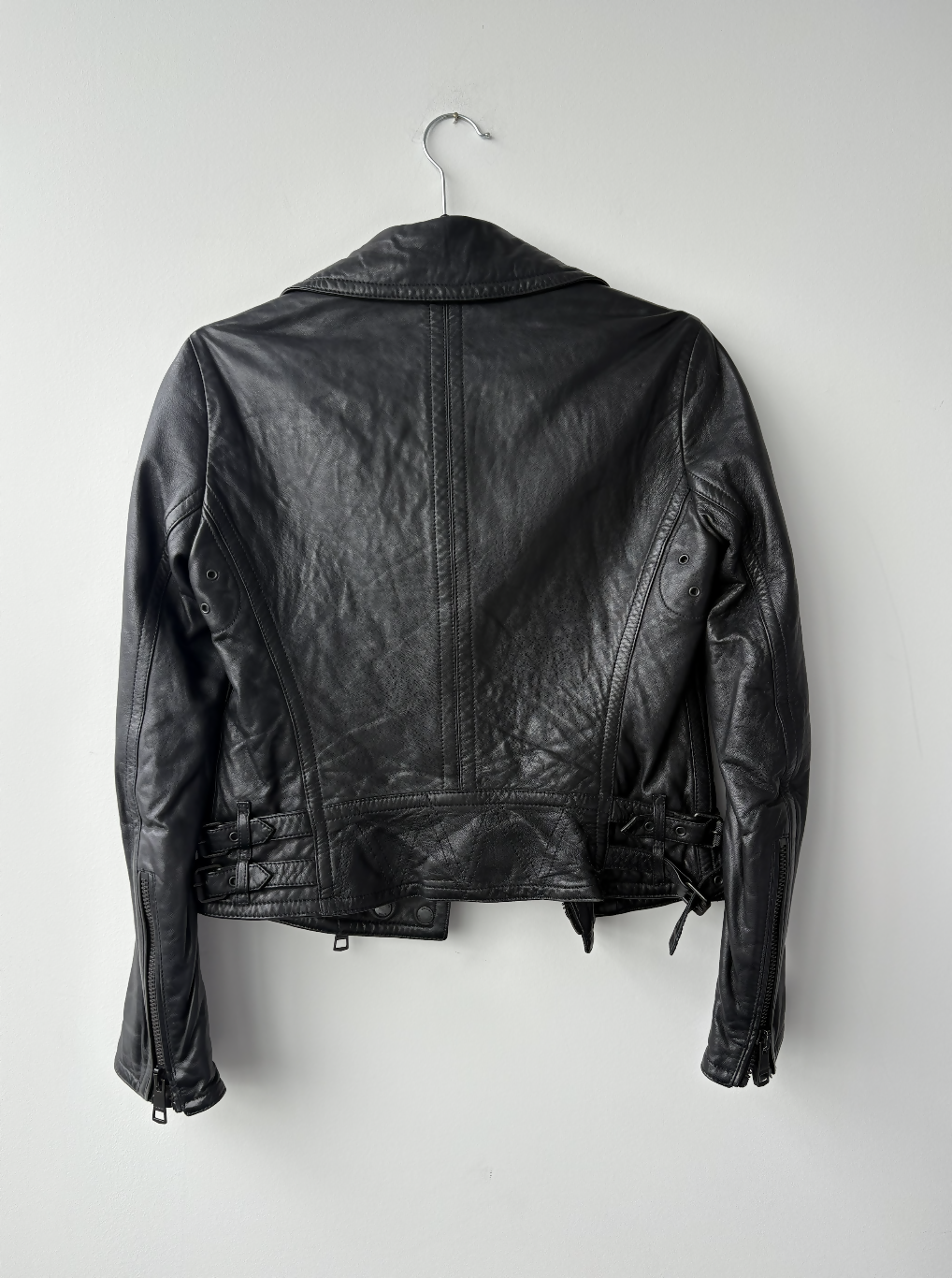The Burberry Brit Black Leather Biker Jacket from EXCHANGE STORE hangs on a metal hanger against a plain white wall, shown from the back, highlighting its moto-inspired stitching and waist buckles.