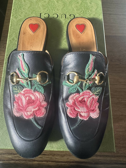 The EXCHANGE Gucci Princetown Black Leather Rose Flower Mule features gold horsebit details and pink rose embroidery, showcased on a patterned green Gucci box.