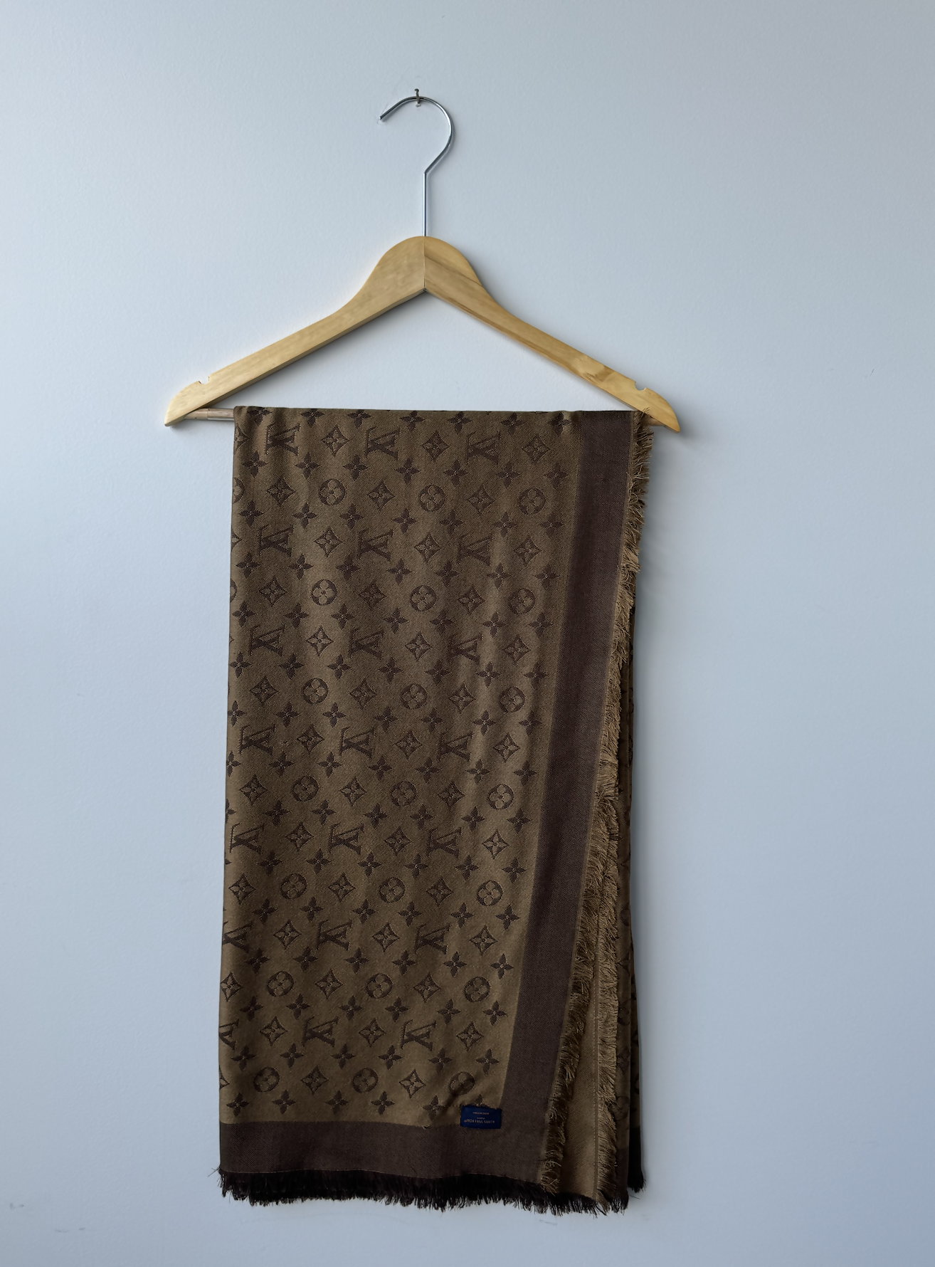 The EXCHANGE Louis Vuitton Scarf, featuring a brown repeating monogram pattern and fringed edges, is neatly draped over a wooden hanger against a plain light gray wall.