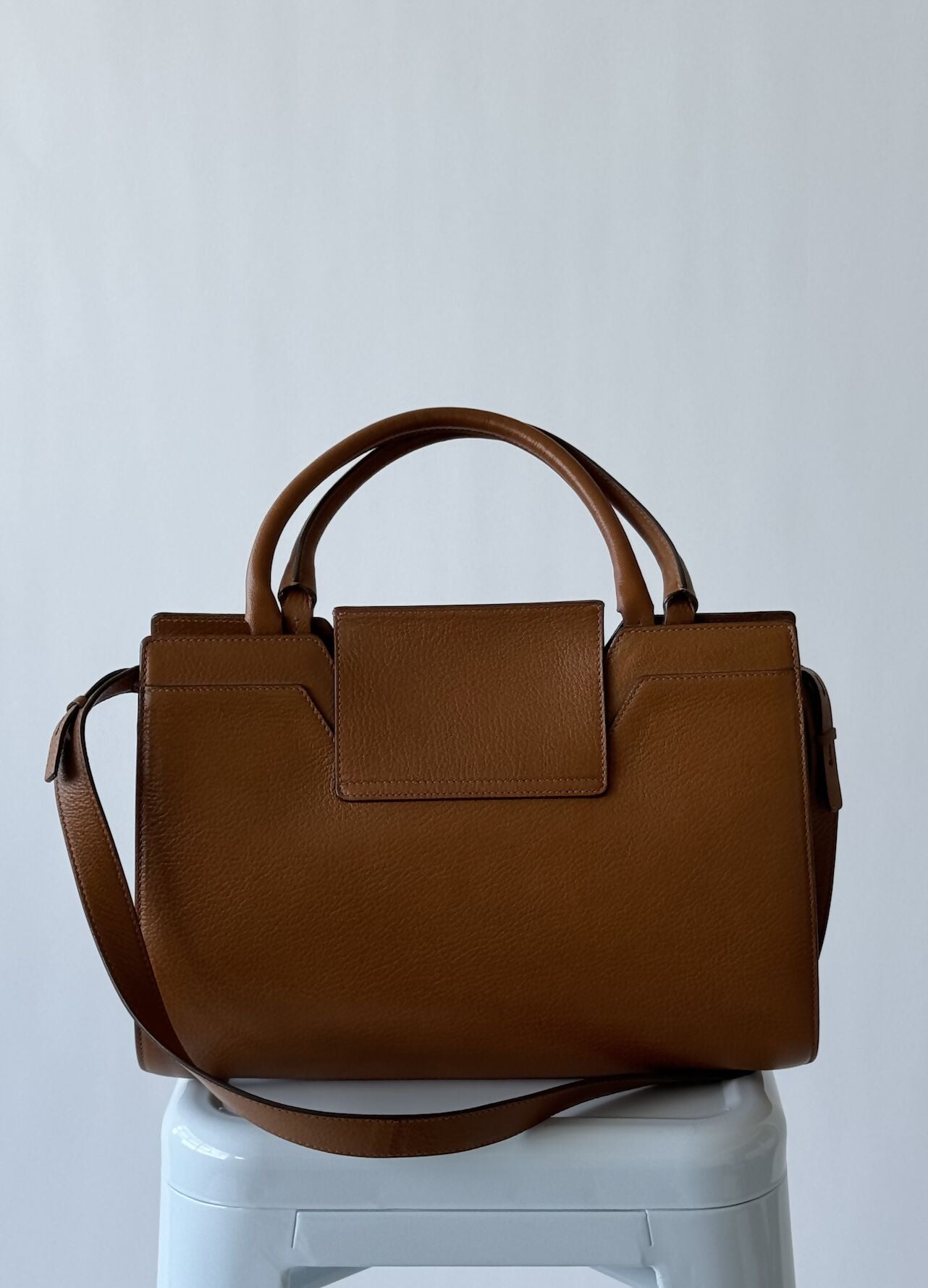 A nutmeg brown EXCHANGE Jimmy Choo - Rebel Tote Bag in Italian goatskin with a top handle, shoulder strap, and front flap closure is displayed on a white stool against a plain light background.
