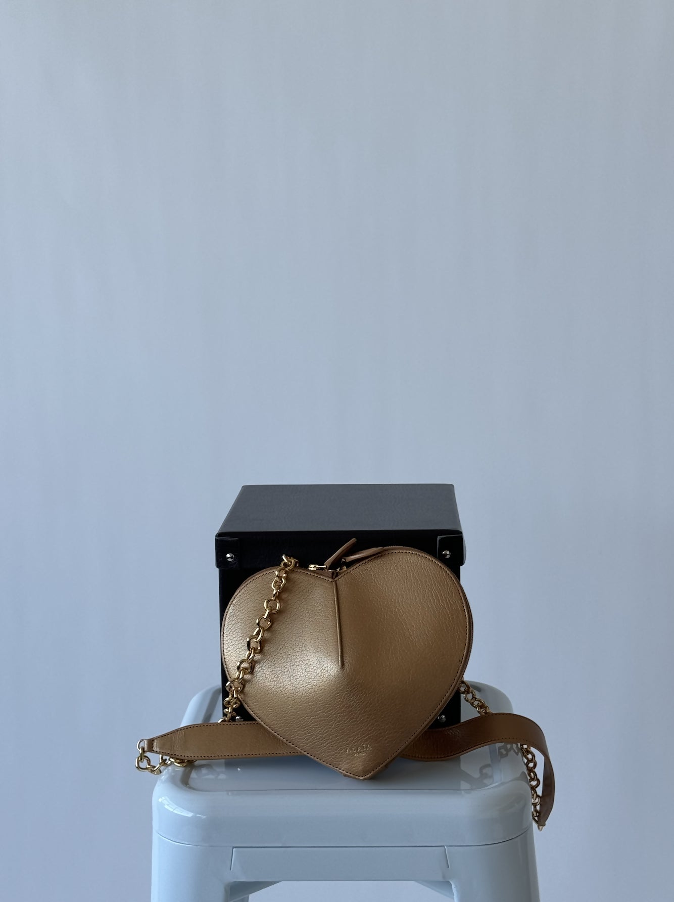 A gold ALAÏA Le Coeur leather shoulder bag by EXCHANGE, presented as a FULL SET with chain strap, rests on a light blue stool in front of a black box against a plain light gray background.