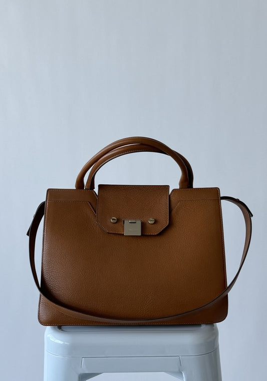 A nutmeg brown EXCHANGE Jimmy Choo - Rebel Tote Bag in Italian goatskin with top handle, shoulder strap, and a silver-clasped front flap sits on a white stool against a plain light gray background.