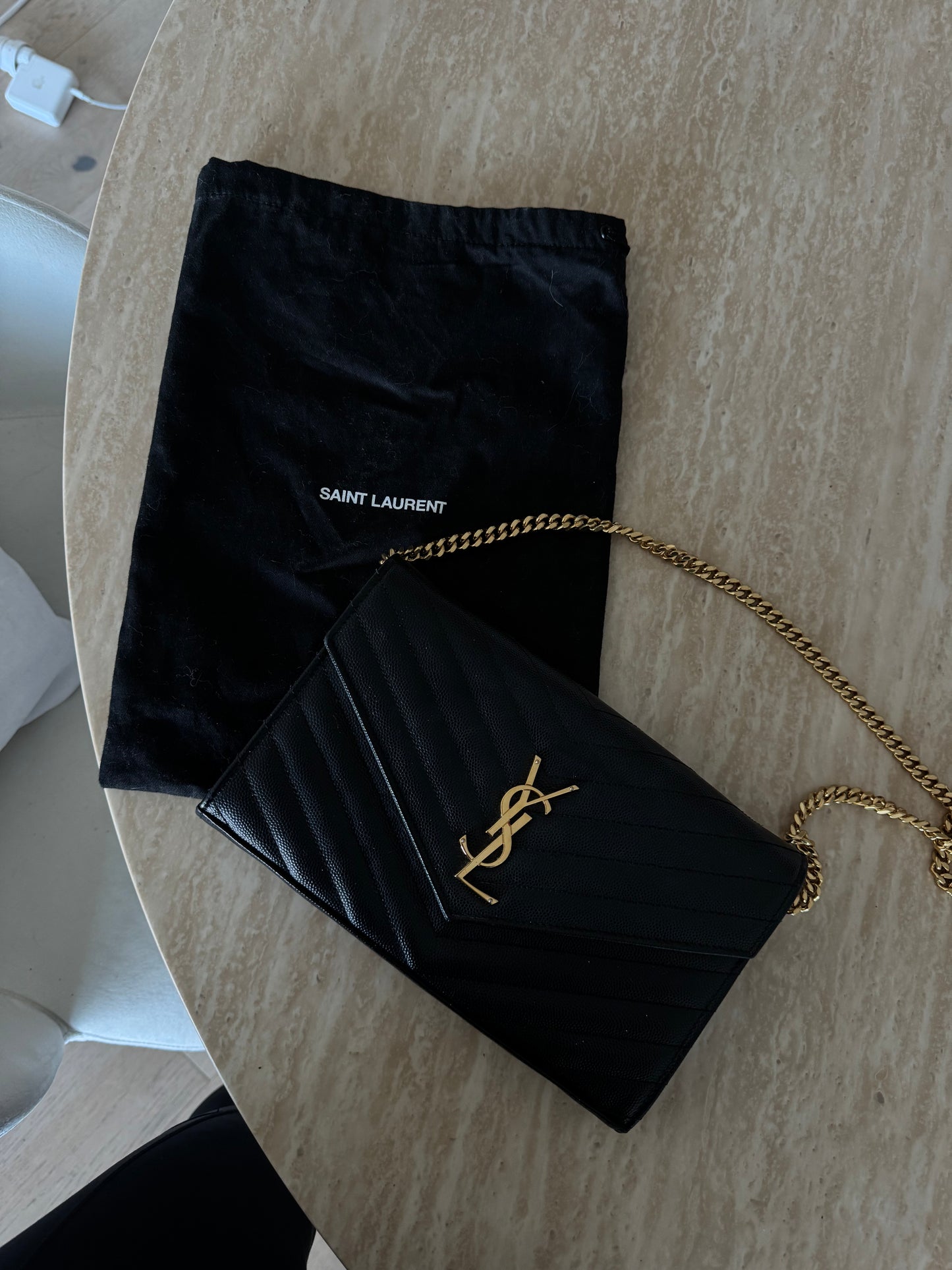 The Saint Laurent Classic Cassandre chain wallet in grain de poudre leather from EXCHANGE STORE, with gold YSL logo and chain, is displayed on a marble surface beside its matching dust bag.