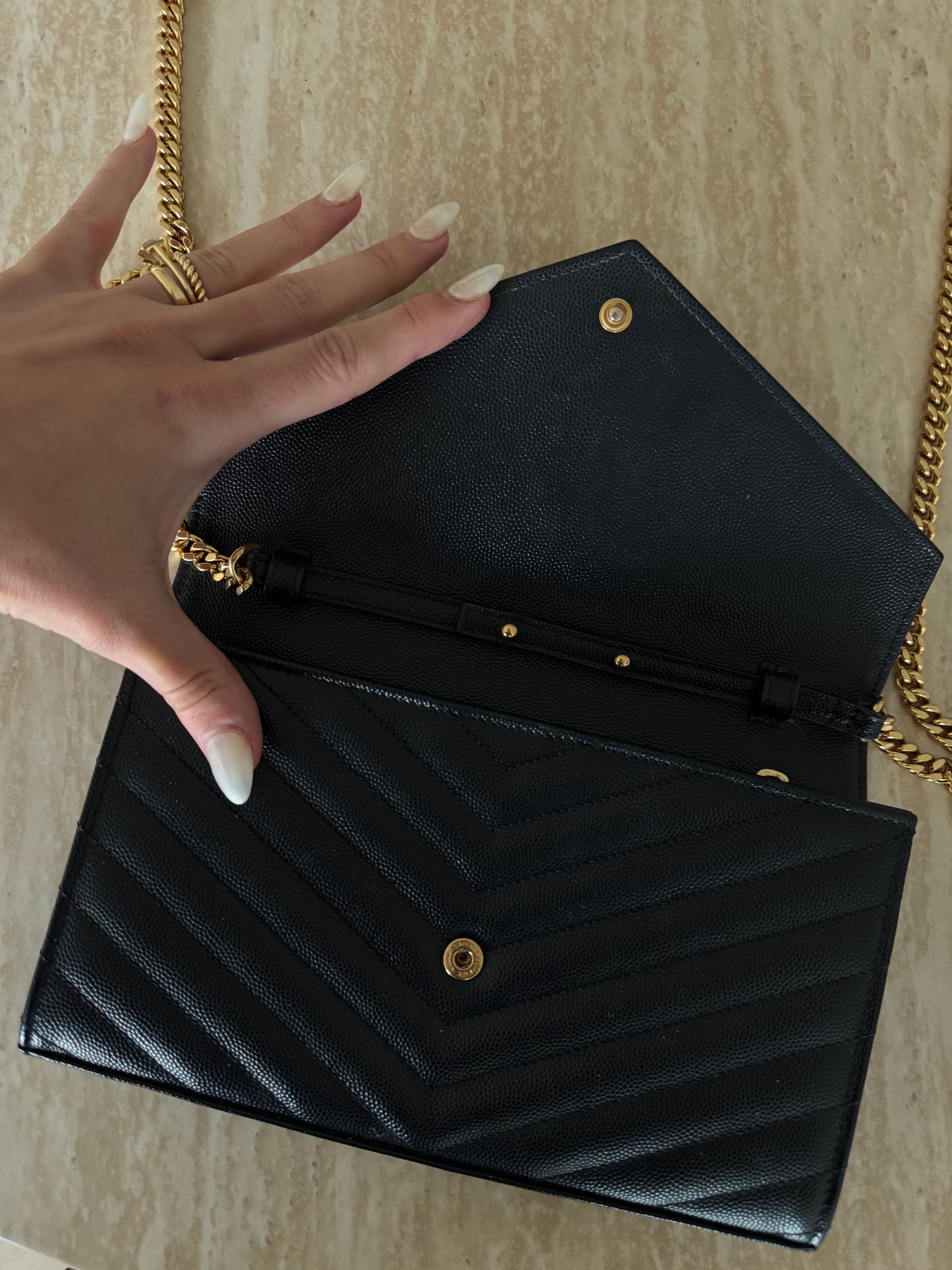 A hand with manicured nails and gold rings opens the EXCHANGE STORE Saint Laurent Classic Cassandre chain wallet in grain de poudre leather on a light marble surface.