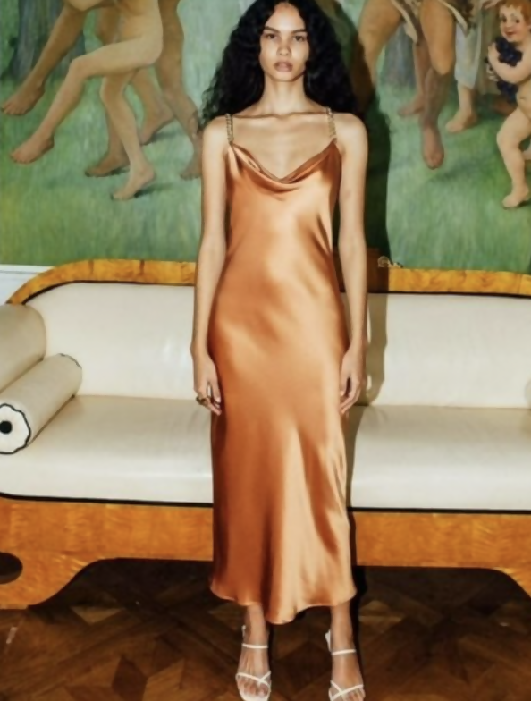 A woman with long curly hair wears the EXCHANGE STORE Olivia Von Halle Bibi Silk Chain Dress and white strappy heels, standing before a beige sofa and a classical painting of cherubs and figures.