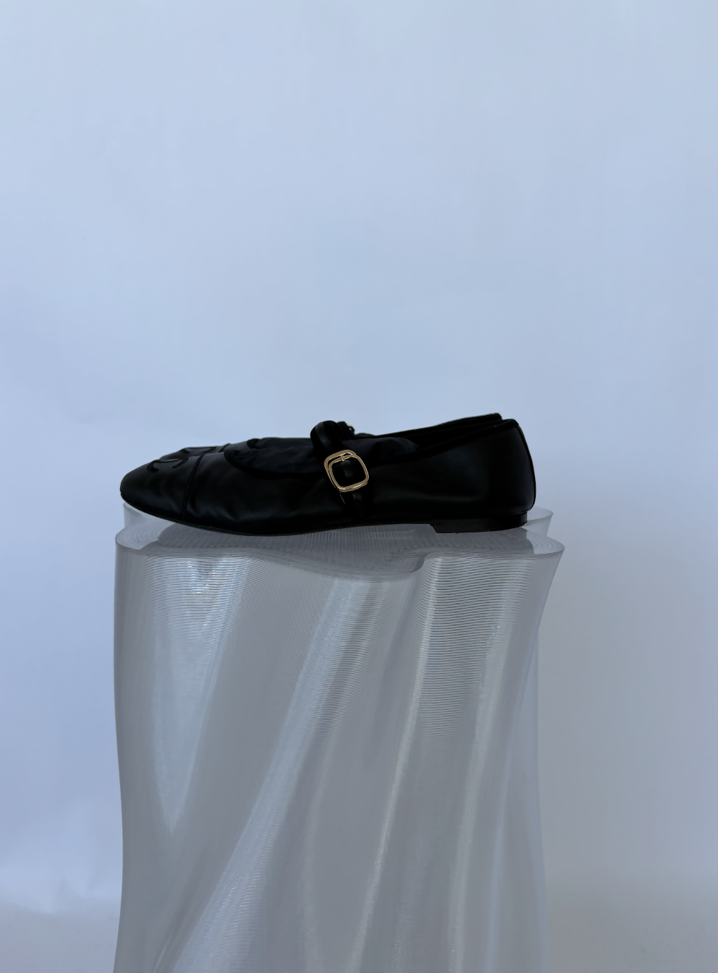A pair of Chanel Mary Jane flats in black lambskin by EXCHANGE STORE, featuring a buckle strap, is displayed on a translucent textured pedestal against a plain light background.