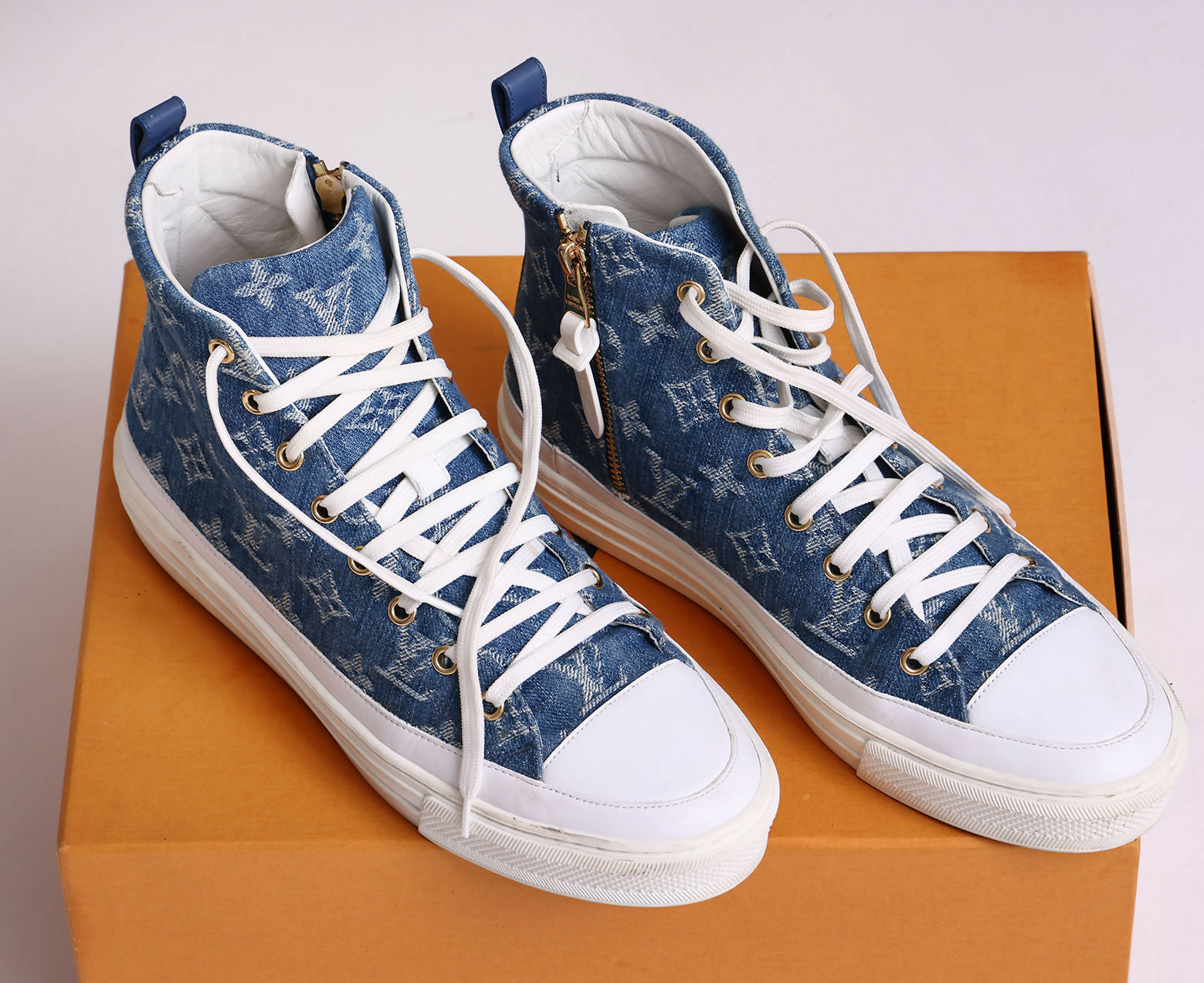 A pair of Louis Vuitton Sneakers by EXCHANGE in denim blue monogram with white laces, white soles, patterned fabric, and gold zippers, displayed on an orange box.