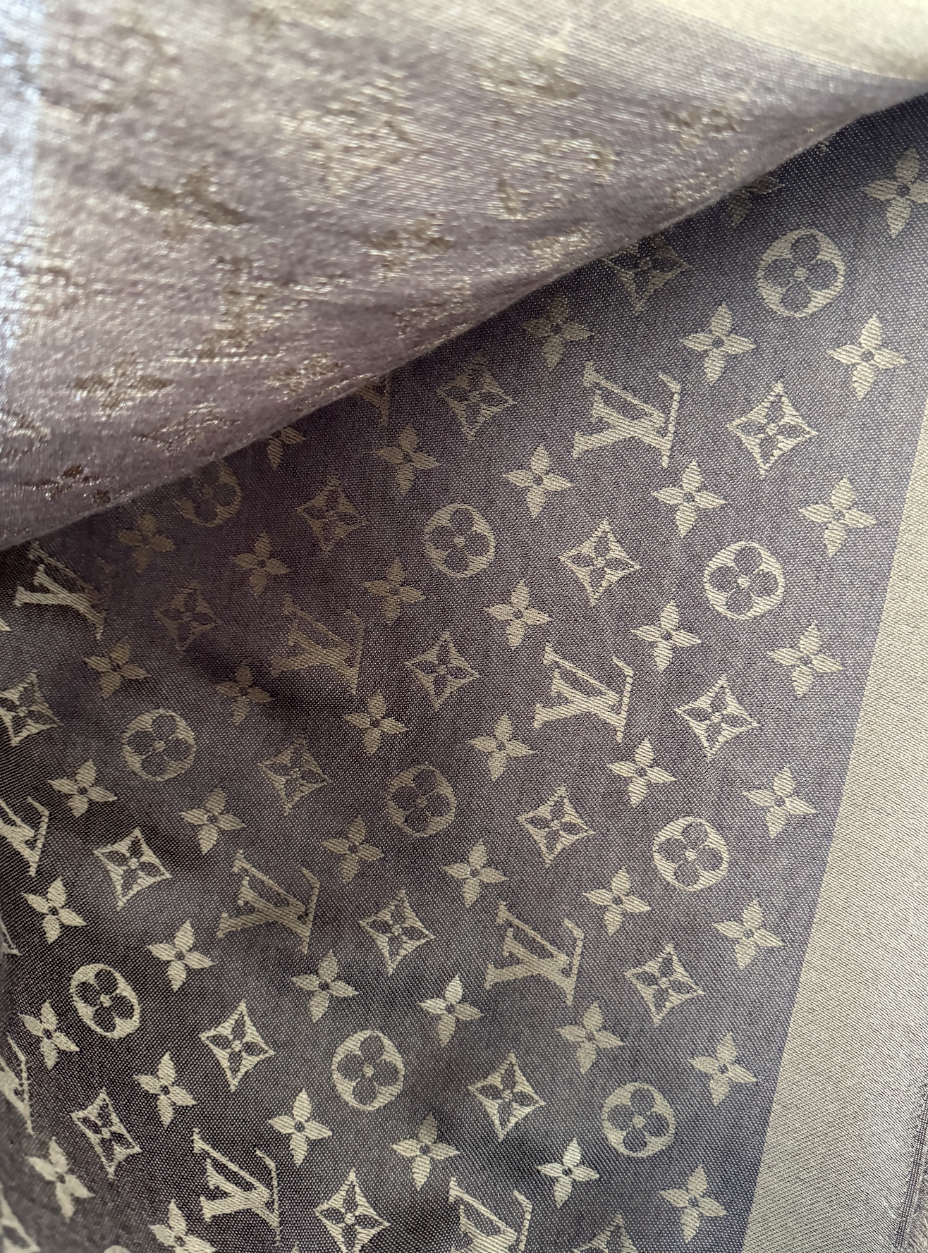 A close-up of the EXCHANGE Louis Vuitton Scarf shows its brown and gold monogram pattern with interlocking LV initials and floral motifs. The soft silk fabric is partially folded, highlighting its luxurious texture.