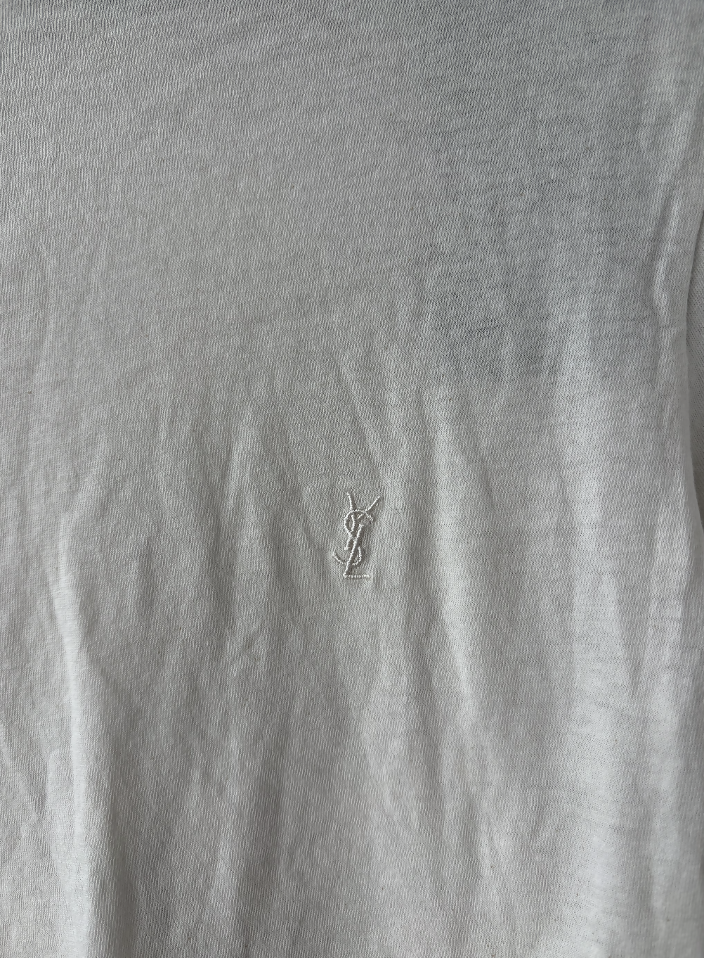 A close-up of the Yves Saint Laurent Beige Logo Cropped T-Shirt by EXCHANGE STORE, showing a small white embroidered YSL logo at the center on soft, beige-grey fabric with visible creases.