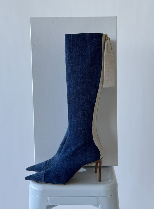 A pair of blue Jacquemus Les bottes Cordao knee-high boots with pointed toes and stiletto heels sits on a white stool. In brand new condition, the boots feature a white zipper at the back that contrasts with the denim texture, all set against a light background to enhance focus.