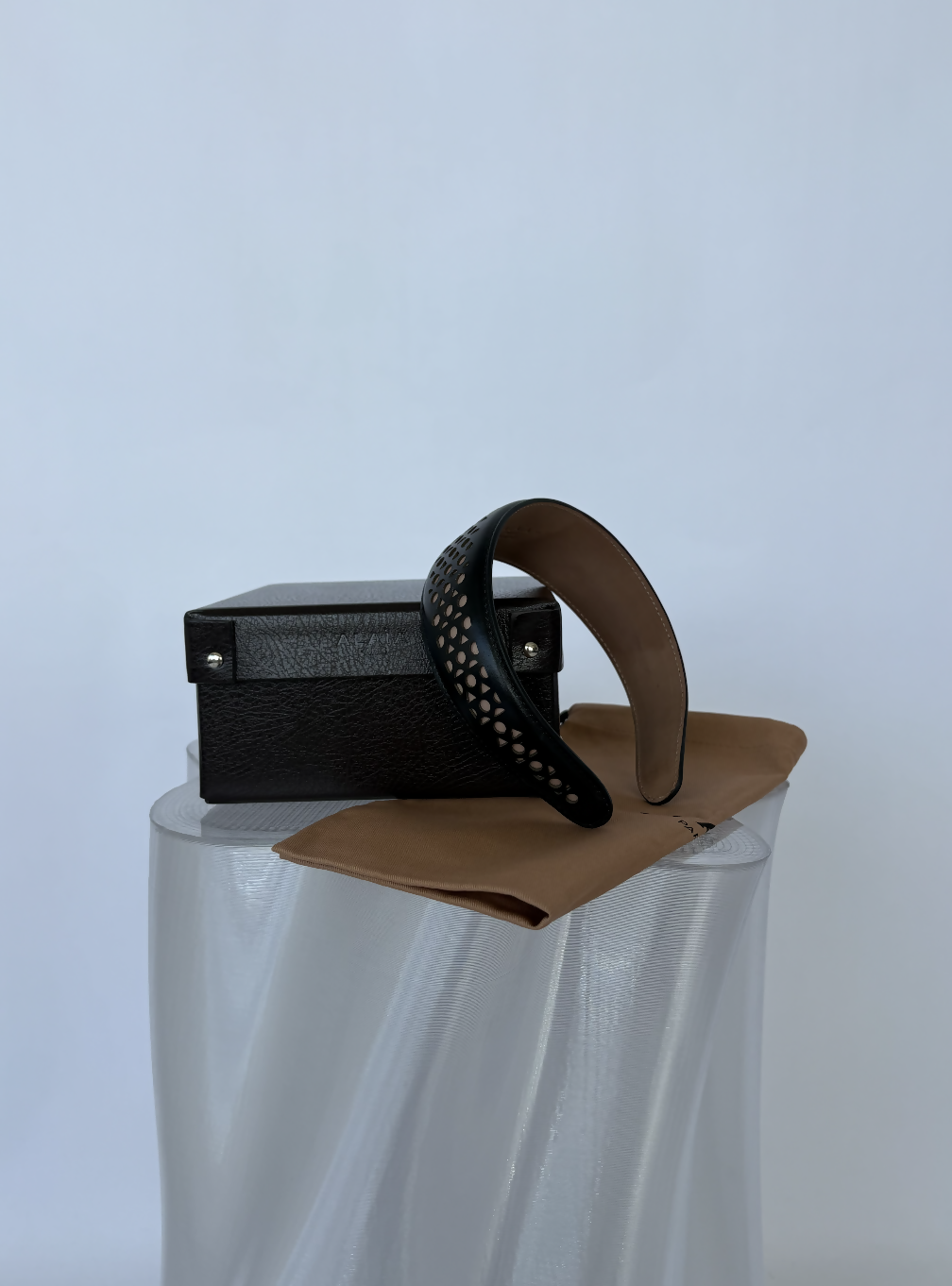 The Alaïa Vienne Laser-Cut Leather Headband - FULL SET by EXCHANGE STORE, in black leather with laser-cut details, is displayed on a light brown cloth before a closed black textured box, all set on a translucent pedestal against a plain white background.