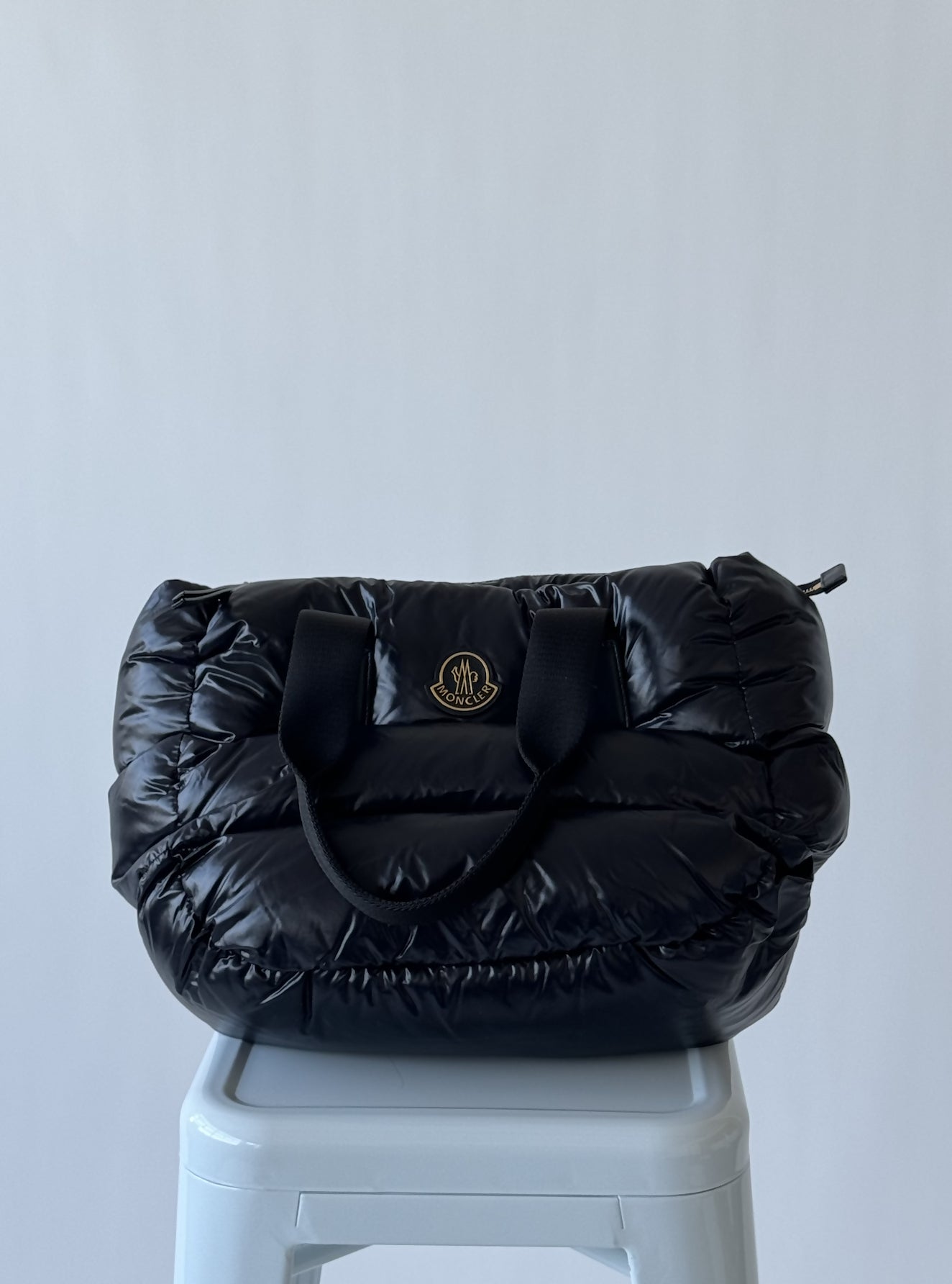 The Moncler Logo Patch Padded Top Handle Bag by EXCHANGE, featuring black puffer-style design, short black handles, and a small gold logo, sits on a light gray plastic stool against a plain light background.