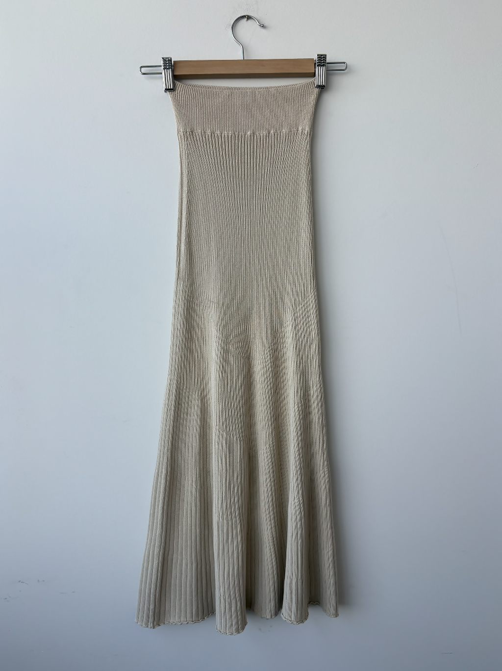 The Dadelszen Beiged Ribbed Knit Skirt by EXCHANGE STORE features a high waistband and flared hem, displayed on a wooden hanger against a white wall—a timeless luxury wardrobe essential.