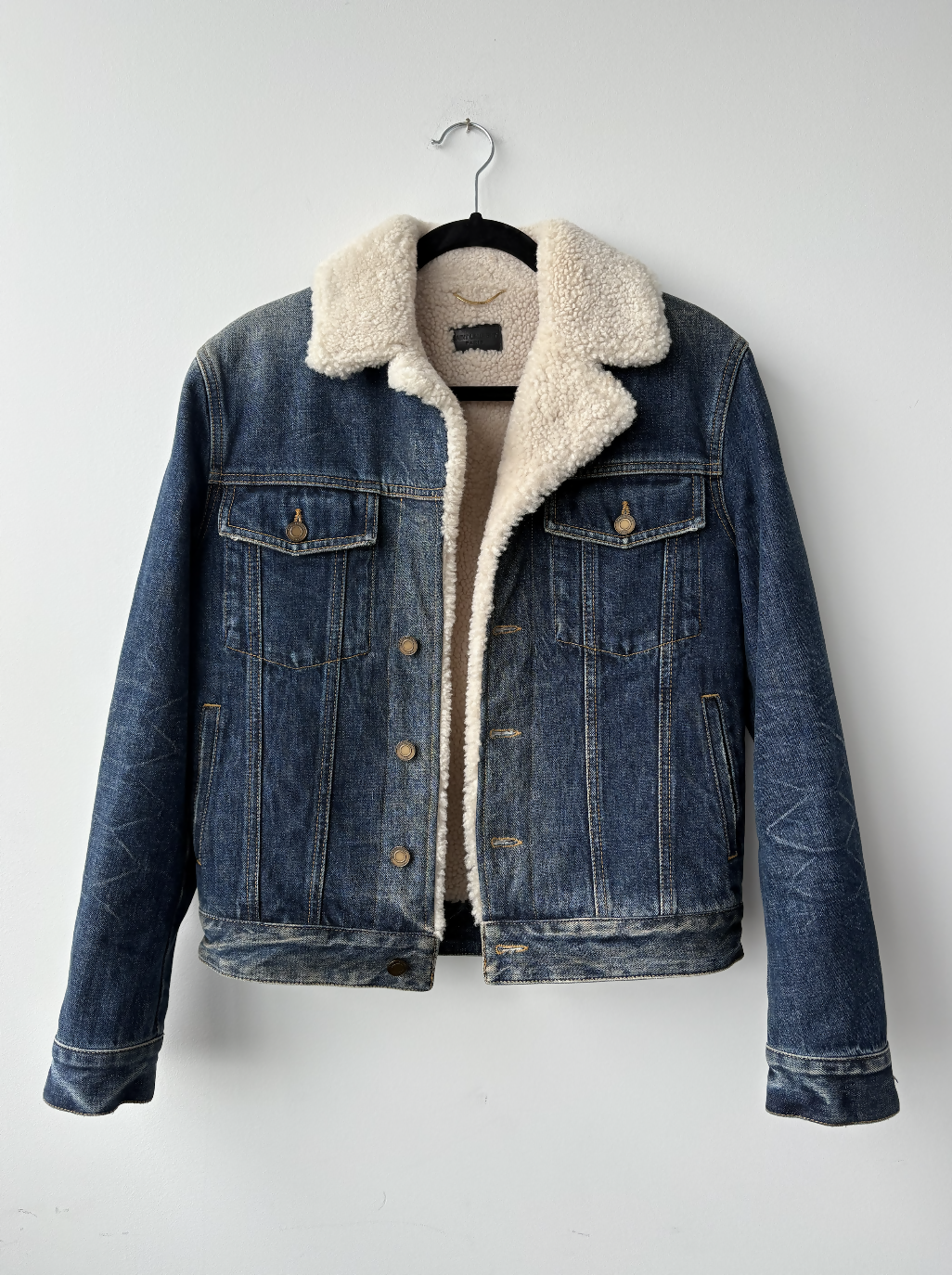 Yves Saint Laurent Paris Denim Shearing Jacket by EXCHANGE STORE, featuring blue denim with white sherpa collar and lining, buttoned front, flap breast pockets, and side pockets, displayed on a black hanger against a white wall.