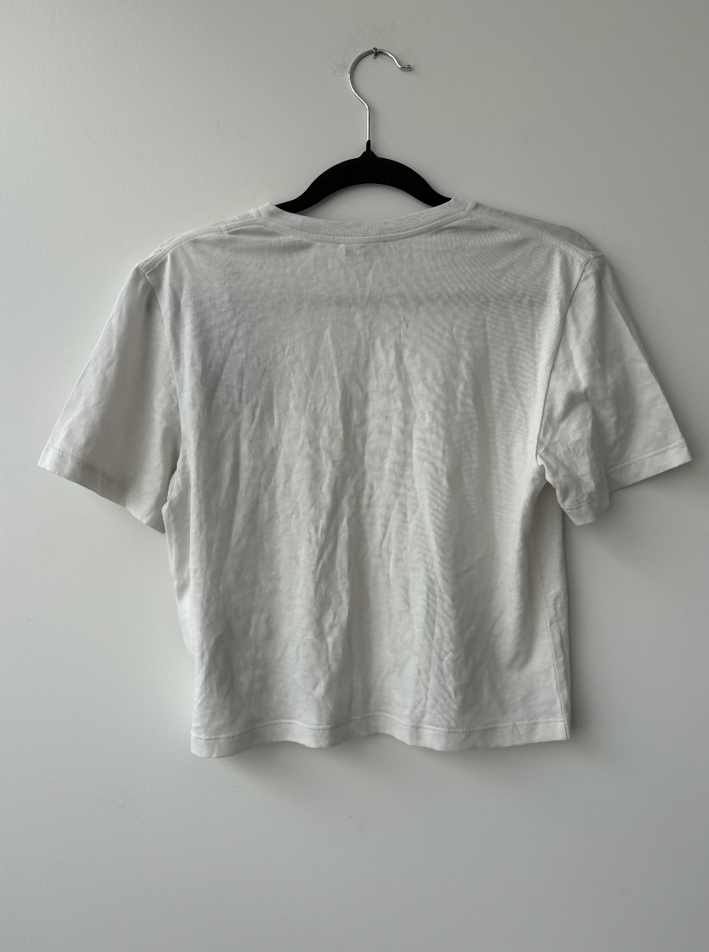 The Yves Saint Laurent Beige Logo Cropped T-Shirt by EXCHANGE STORE hangs on a black hanger against a white wall, displayed with the back facing forward. The shirt is plain, beige-grey, and appears slightly wrinkled.
