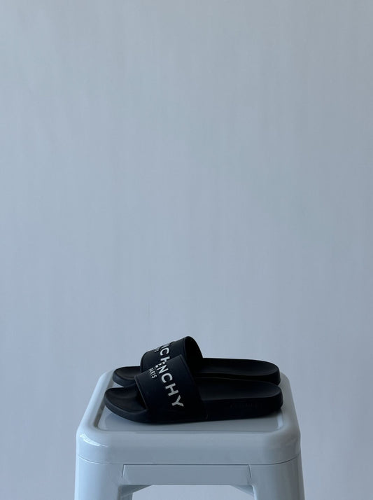 A pair of black Givenchy Logo Detailed Open Toe Slides by EXCHANGE in size 39, featuring white lettering, rests on a white stool against a light gray background. Available now at Faraday's.