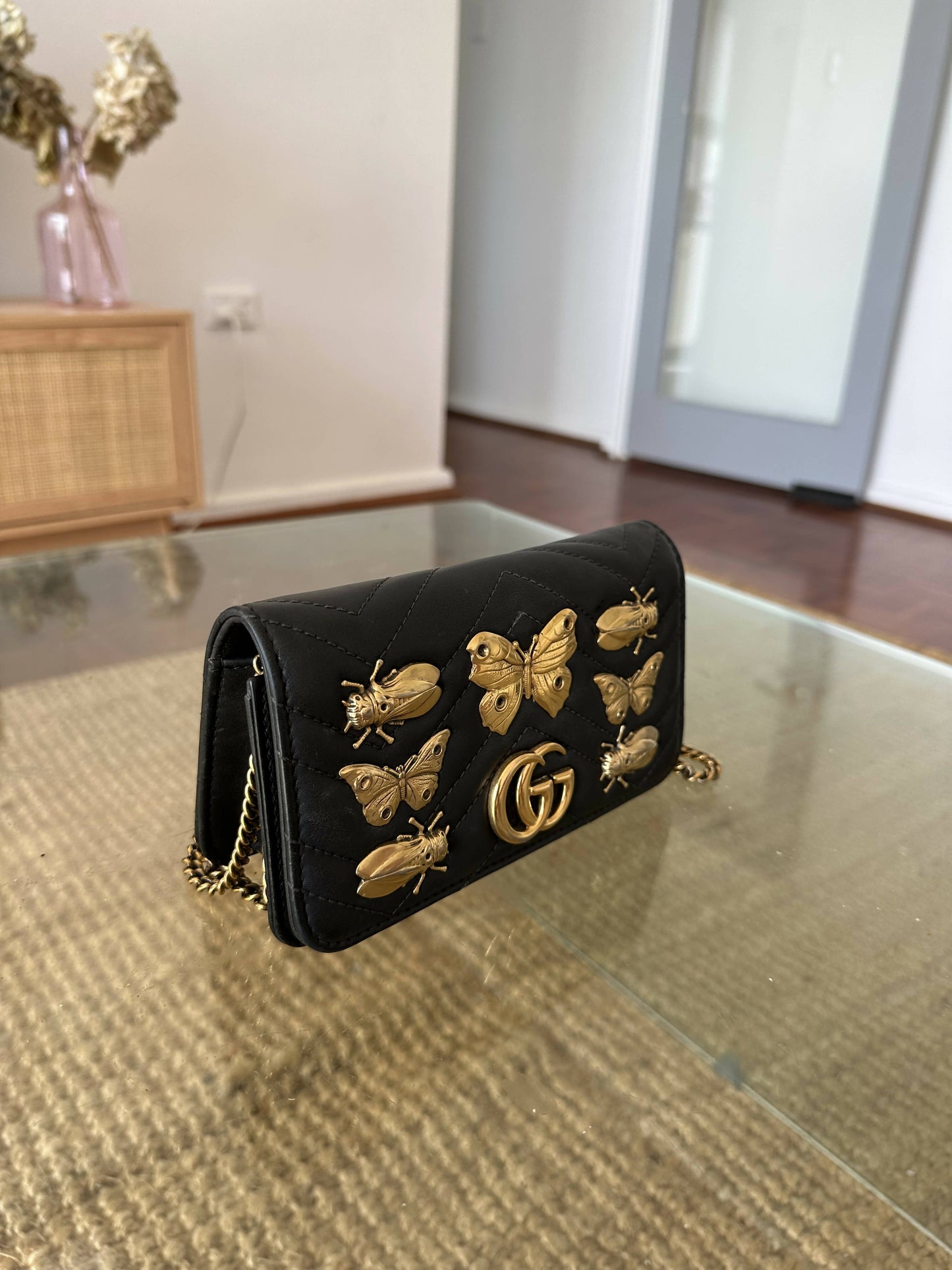 A black EXCHANGE Gucci Marmont Butterfly Bag with gold studs and a chain strap rests on a woven surface in a modern living room.