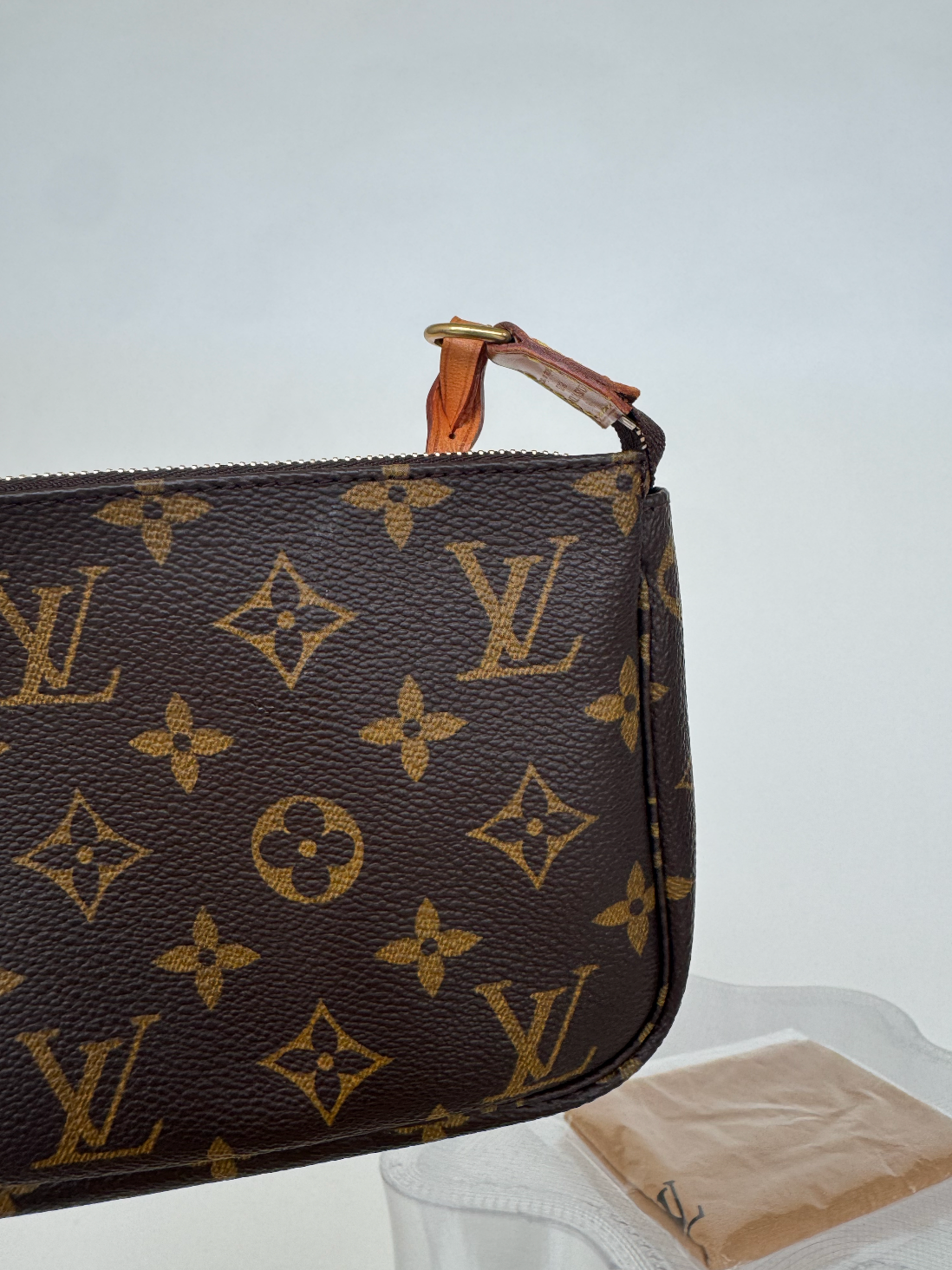 A close-up of the Louis Vuitton - Monogram Pochette by EXCHANGE STORE, showing gold LV and floral patterns, a leather strap, and zipper, in perfect condition with date code visible against a plain light background.