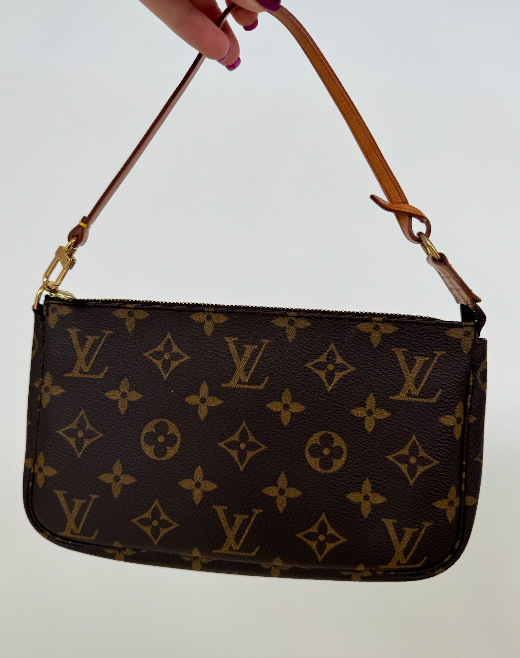 A hand holds the EXCHANGE STORE Louis Vuitton - Monogram Pochette, featuring the classic LV monogram and brown leather strap, against a white background. In perfect condition, it comes complete with its original dust bag.