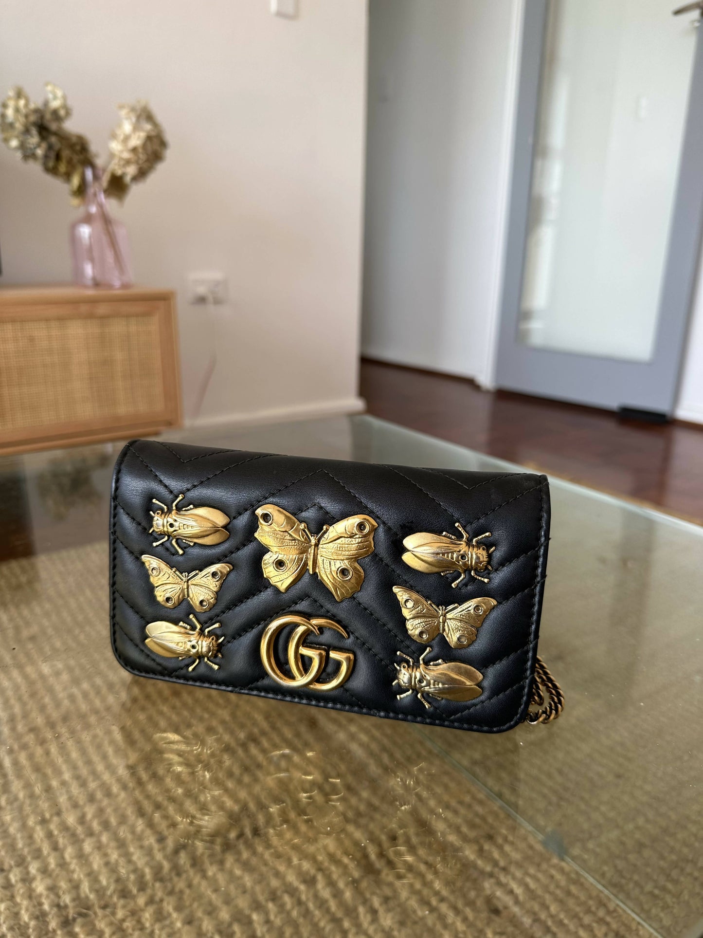A black Gucci Marmont Butterfly Bag by EXCHANGE, featuring quilted calfskin, gold GG logo, and butterfly and bee studs, sits on a glass table in a modern living room.