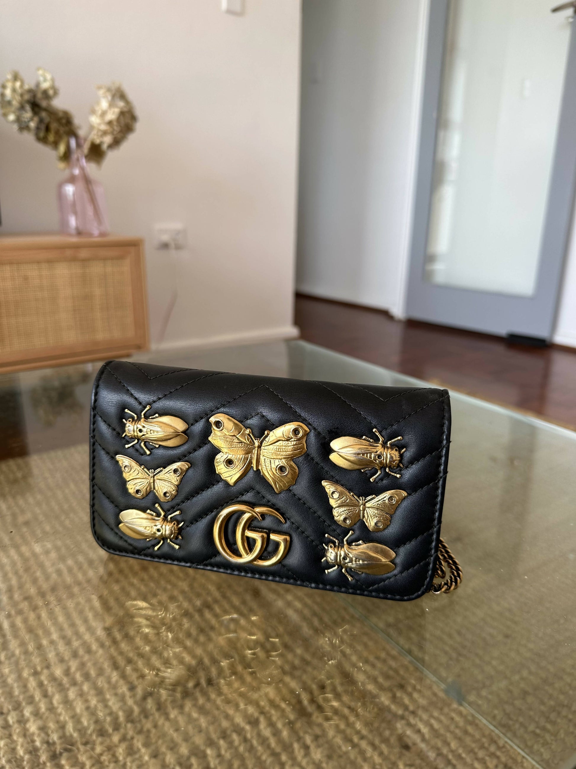A black Gucci Marmont Butterfly Bag by EXCHANGE, featuring quilted calfskin, gold GG logo, and butterfly and bee studs, sits on a glass table in a modern living room.