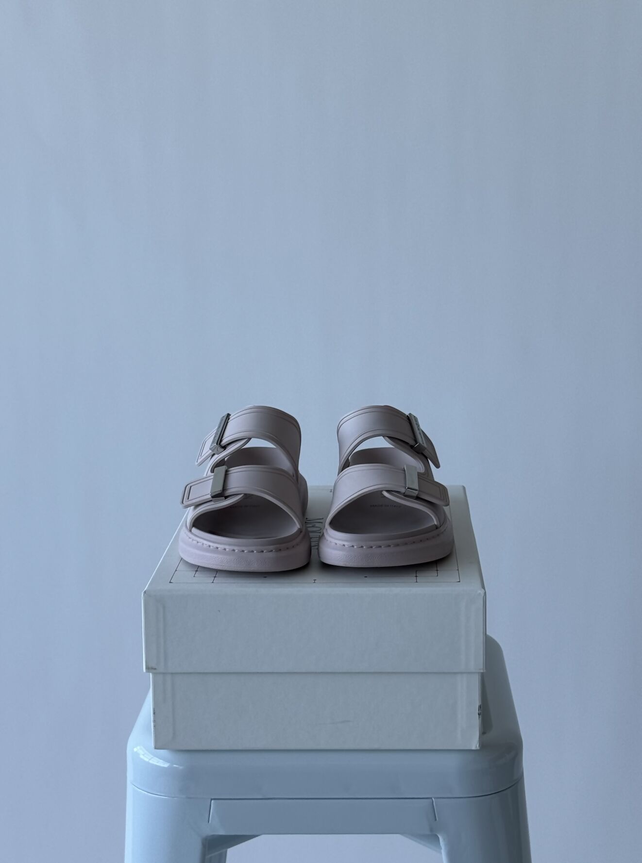 The Alexander McQueen Women Hybrid slide pink/silver by EXCHANGE in size 37 are displayed side by side atop a closed white shoebox, which rests on a light blue stool against a plain light gray background.