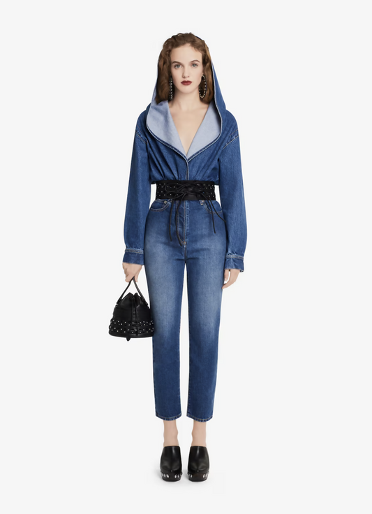 A woman faces forward in a denim hooded jacket cinched with a wide black corset belt, wearing EXCHANGE ALAÏA High Waist Pants, black platform clogs, large earrings, and holding a small black studded handbag.