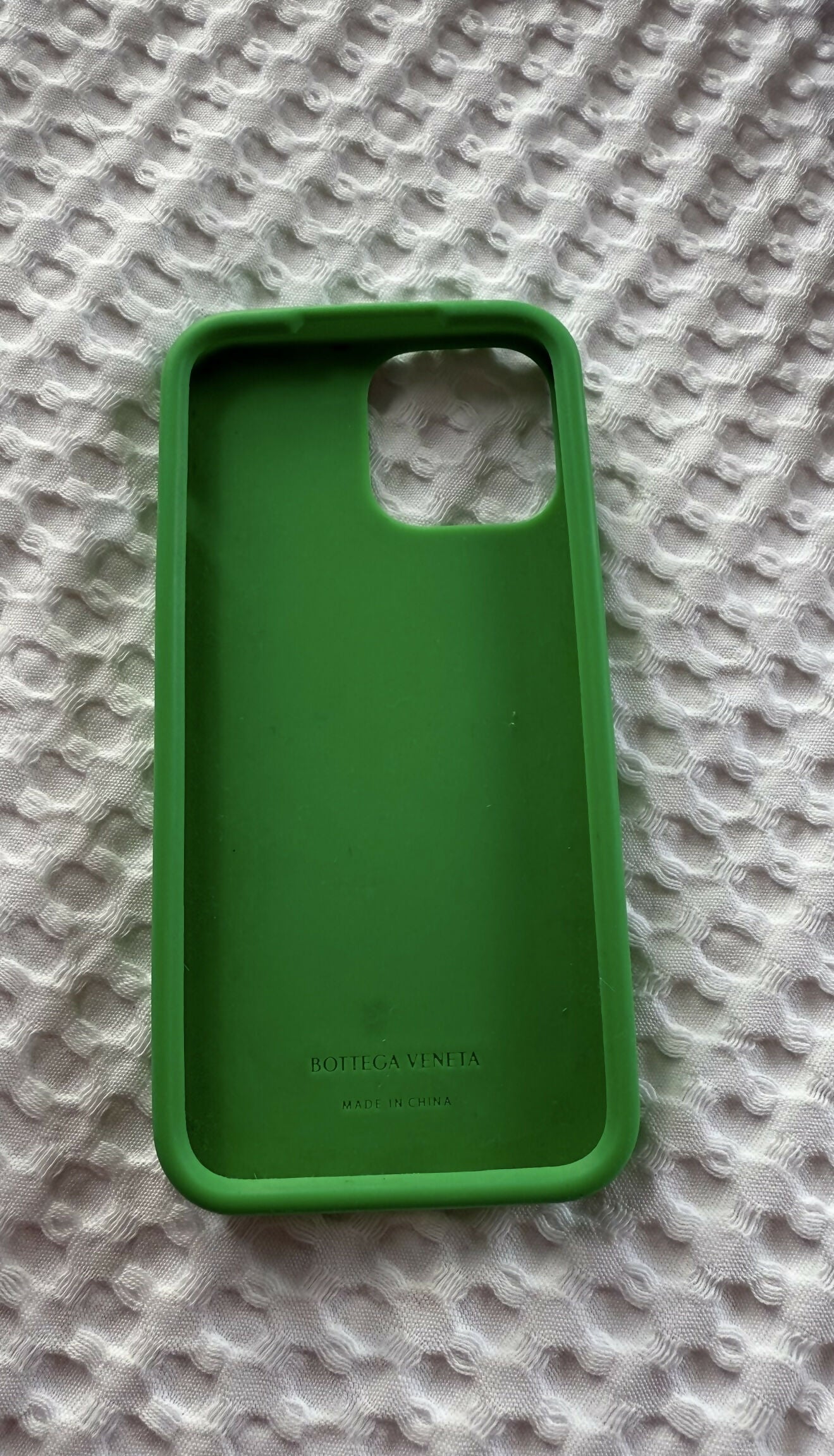 An Intrecciato Rubber iPhone 13 Pro Max Case by EXCHANGE STORE with a camera cutout rests on textured white fabric. The stylish case features the brand name and Made in China printed inside.