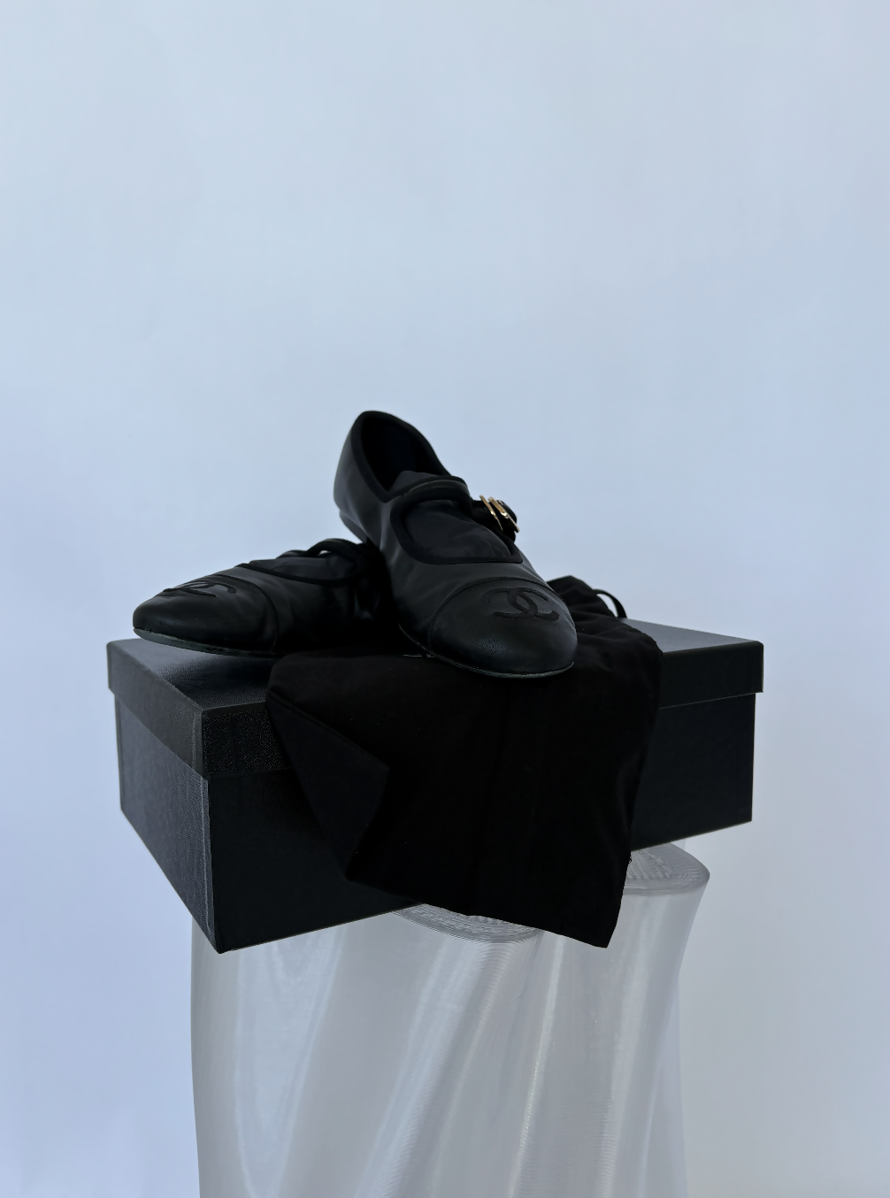 The Chanel Mary Jane flats in Lambskin - FULL SET by EXCHANGE STORE rest atop a black box, partly covered with a black cloth, all arranged on a translucent pedestal—a sleek and collectible display.