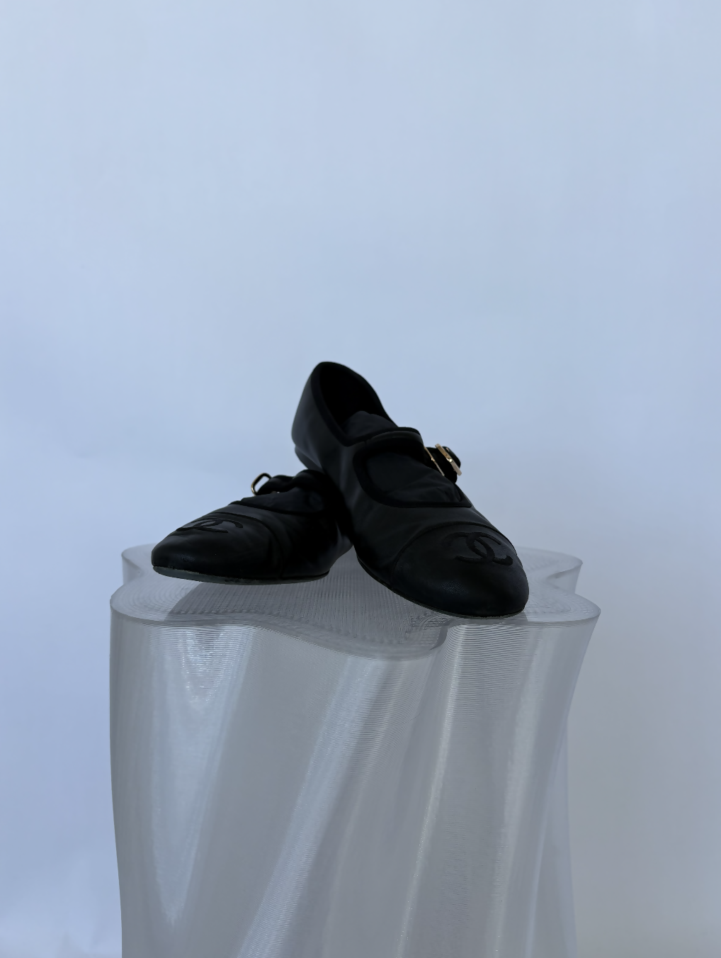 A pair of Chanel Mary Jane flats in lambskin by EXCHANGE STORE—with a gold buckle—rests on a wavy-edged translucent pedestal against a white background, making it a must-have full set for collectors.
