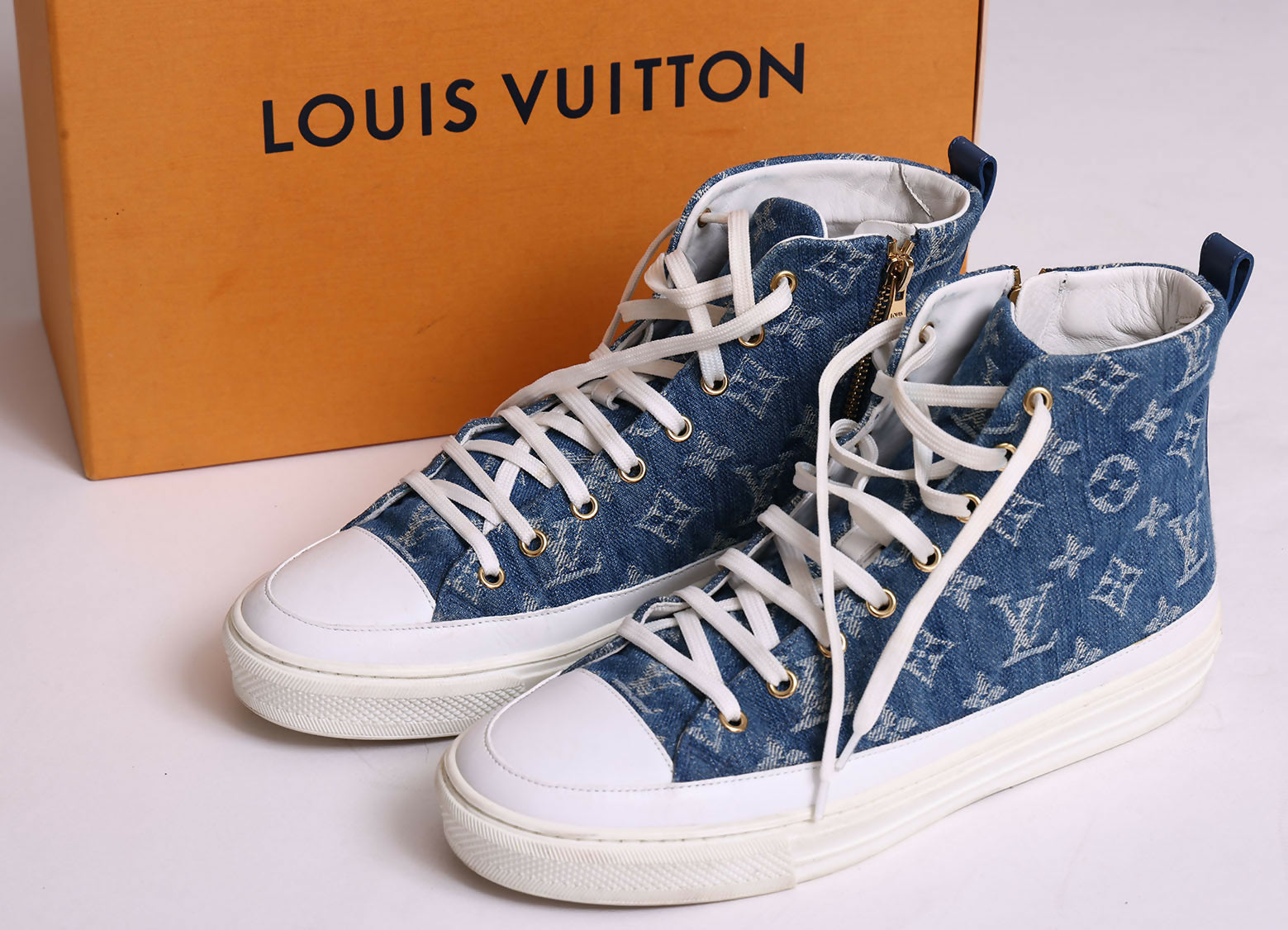 A pair of Louis Vuitton Sneakers by EXCHANGE in denim blue monogram with white soles, laces, and toe caps, shown in front of an orange Louis Vuitton box.