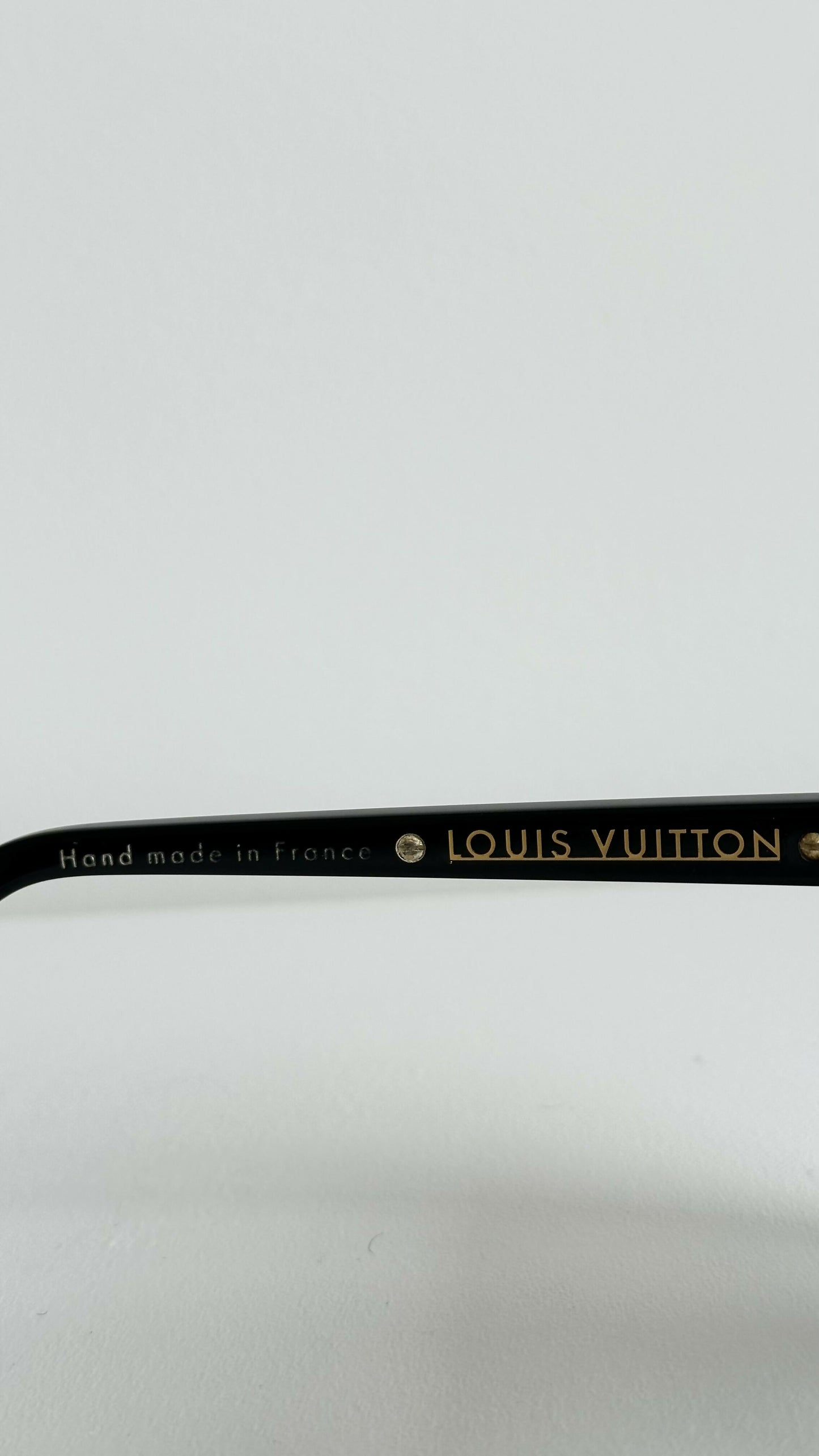 Close-up of the black arm of Louis Vuitton Evidence Noir W sunglasses by EXCHANGE STORE, showing gold "LOUIS VUITTON" lettering and "Hand made in France" against a plain white background.