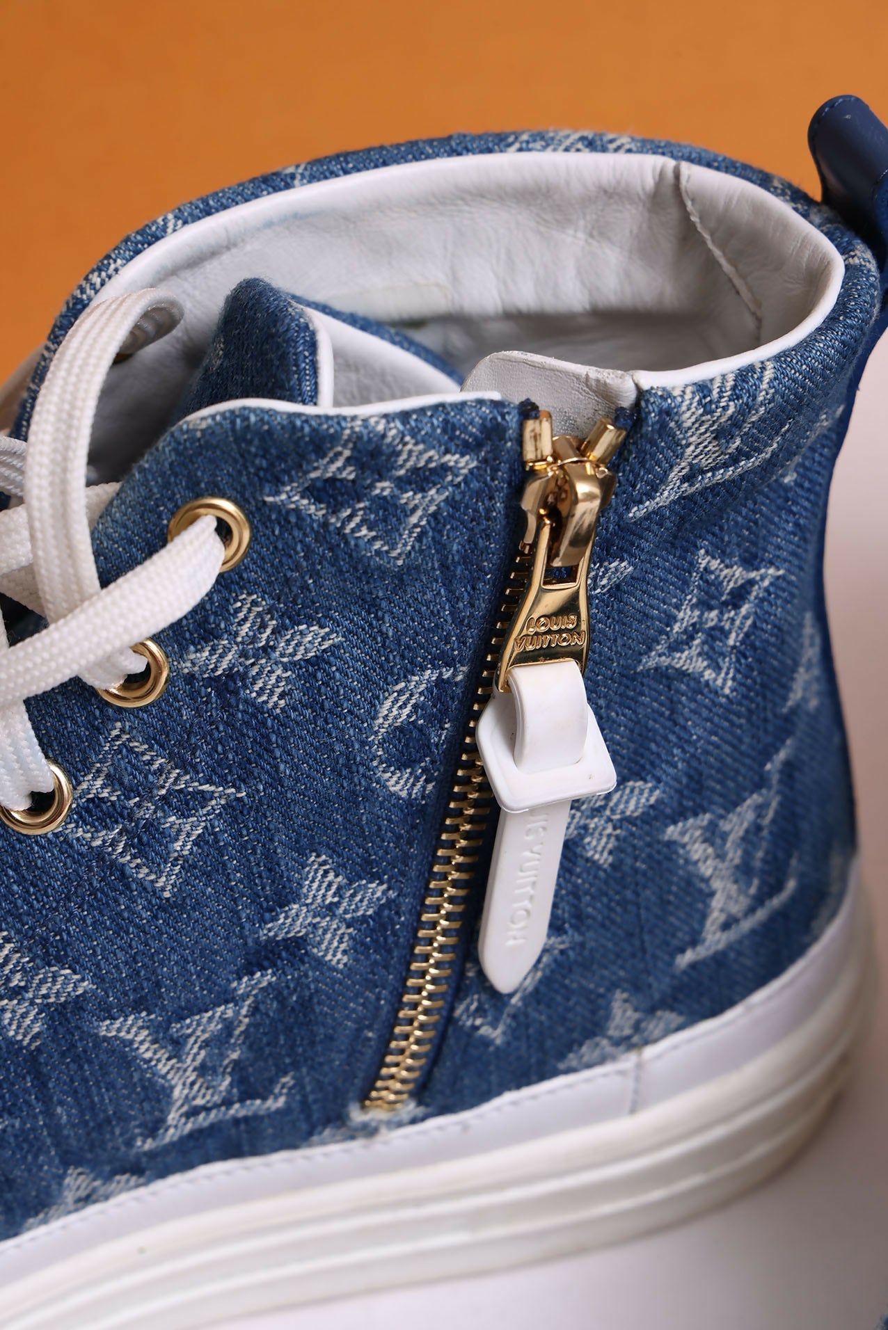Close-up of EXCHANGE Louis Vuitton Sneakers in denim blue monogram, featuring white laces, a gold zipper with a white pull tab, and a white sole set against an orange background.