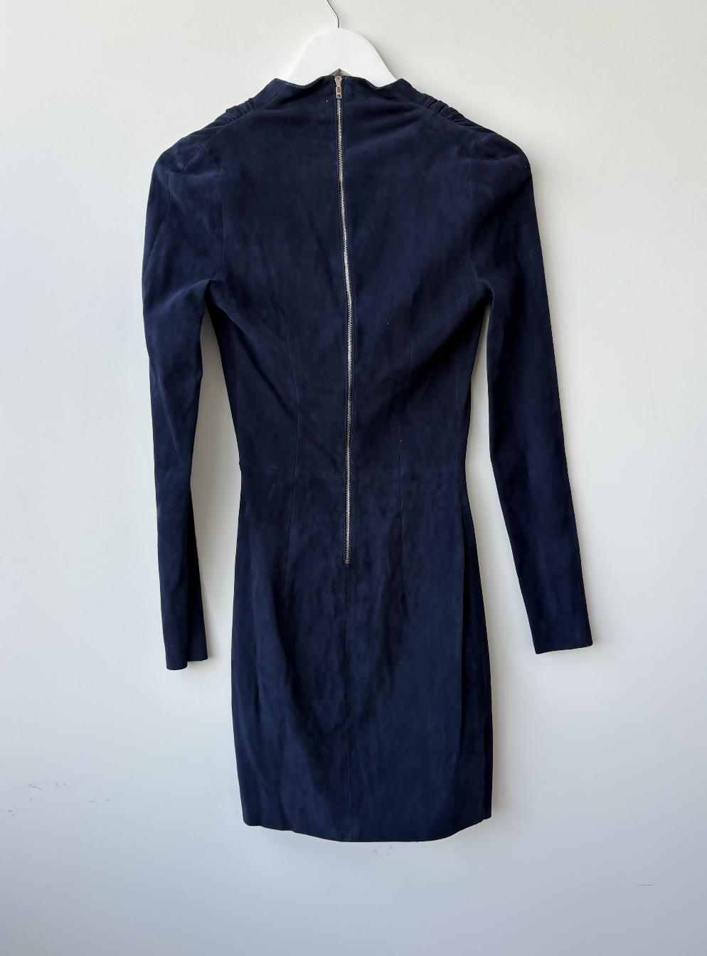The EXCHANGE STORE Jitrois Navy Velvet Longsleeve Mini Dress hangs on a white hanger against a light wall, featuring a fitted silhouette, high neckline, and visible gold back zipper for a subtle rock-chic look.
