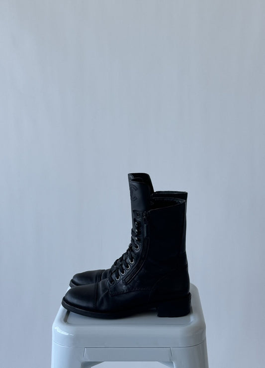 The EXCHANGE CHANEL Sheepskin Quilted CC Tall Lace Up Combat Boots - Vintage, in black leather with side zippers, are displayed on a white stool against a plain light gray background.