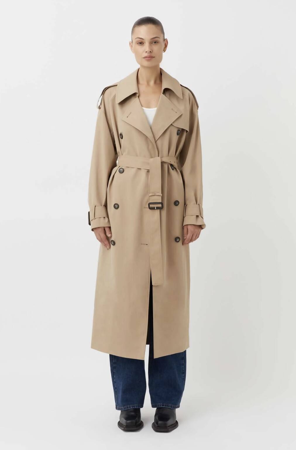 A person faces forward against a plain white background, wearing an EXCHANGE Evans Trench Coat Camilla & Marc in beige (extra small, worn once, great condition) with a belt, paired with blue jeans, a white top, and black shoes.