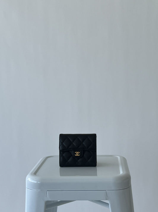 An EXCHANGE CHANEL Caviar Quilted Compact Flap Wallet with a gold logo sits on a light gray plastic stool against a plain, light background.