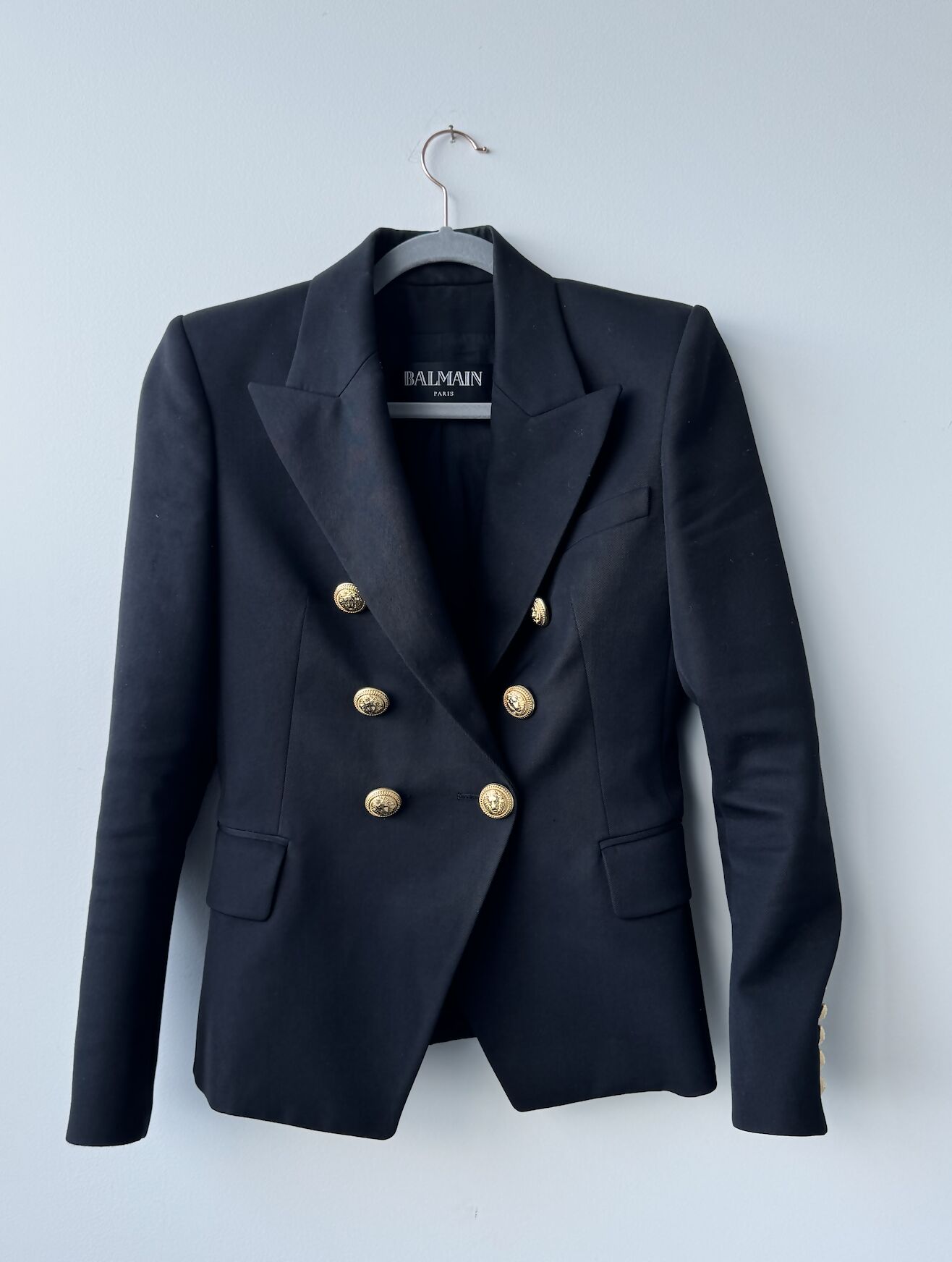 A Balmain Blazer Black by EXCHANGE in size 38 hangs on a hanger, showcasing gold buttons, structured shoulders, and notched lapels against a light-colored wall.