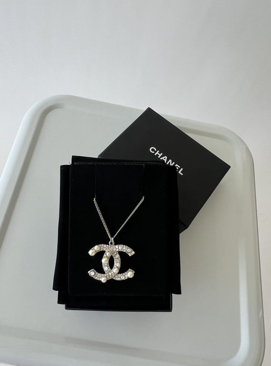 The CHANEL Crystal Pearl CC Pendant Necklace Silver by EXCHANGE, featuring a crystal-studded logo, rests on black velvet beside a partial Chanel box and Certificate of Authenticity atop a light gray surface.